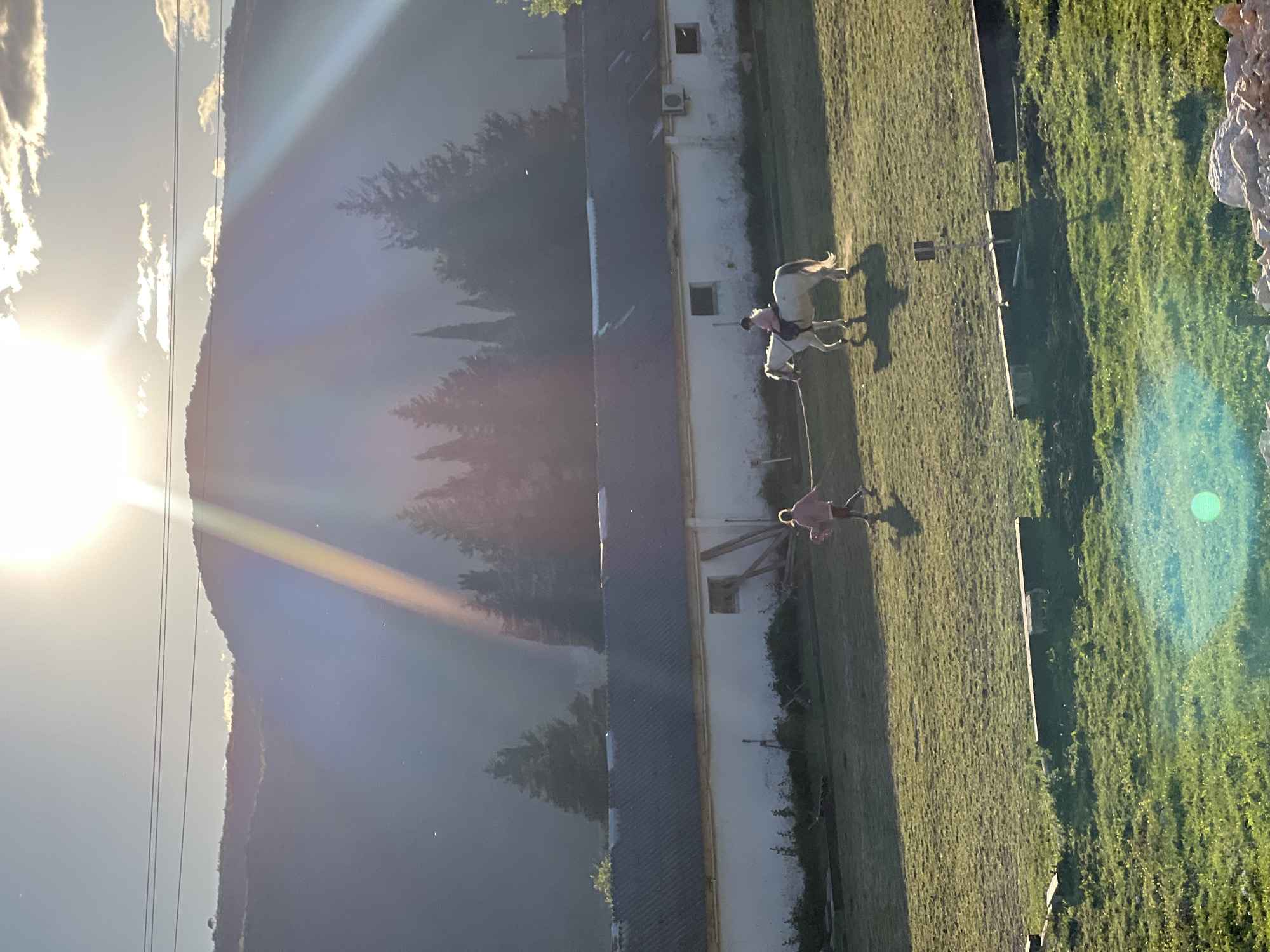 The image shows a rural scene in Podlastva (Poljice), Montenegro. A white building with a dark roof runs horizontally through the center of the frame. To the left is a mountain range against a bright sky with the sun visible. To the right of the building, a grassy field is visible. In the field, a person is leading a light-colored horse. Another person appears to be riding the horse.