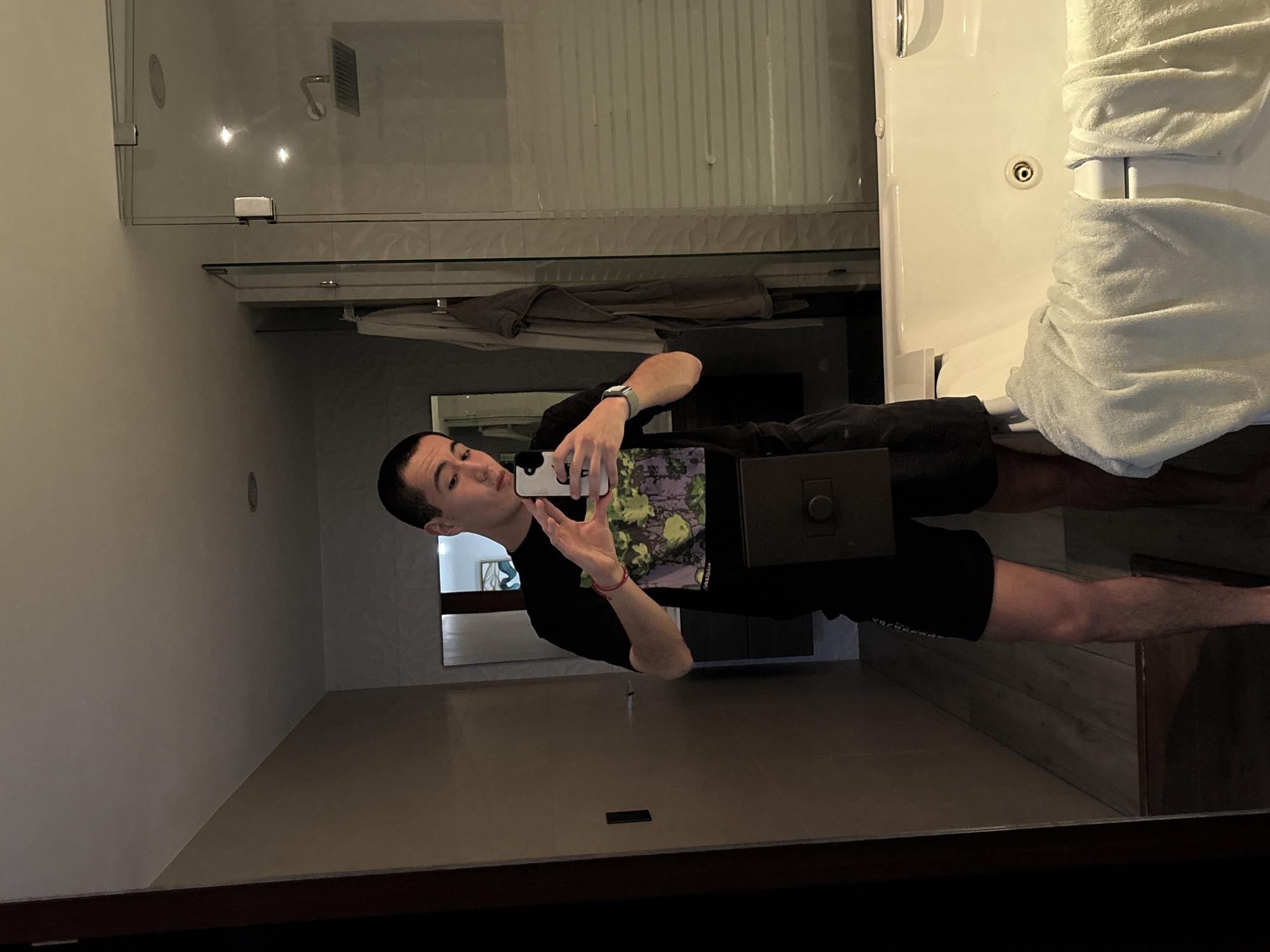A man is taking a picture of himself in a bathroom mirror. He is wearing a black t-shirt with a graphic on the front and black shorts. He is holding a cell phone in his right hand, which he is using to take the picture. The bathroom has a shower with a glass door, a white sink, and white towels. The location is San Bernardino Tlaxcalancingo, Mexico.