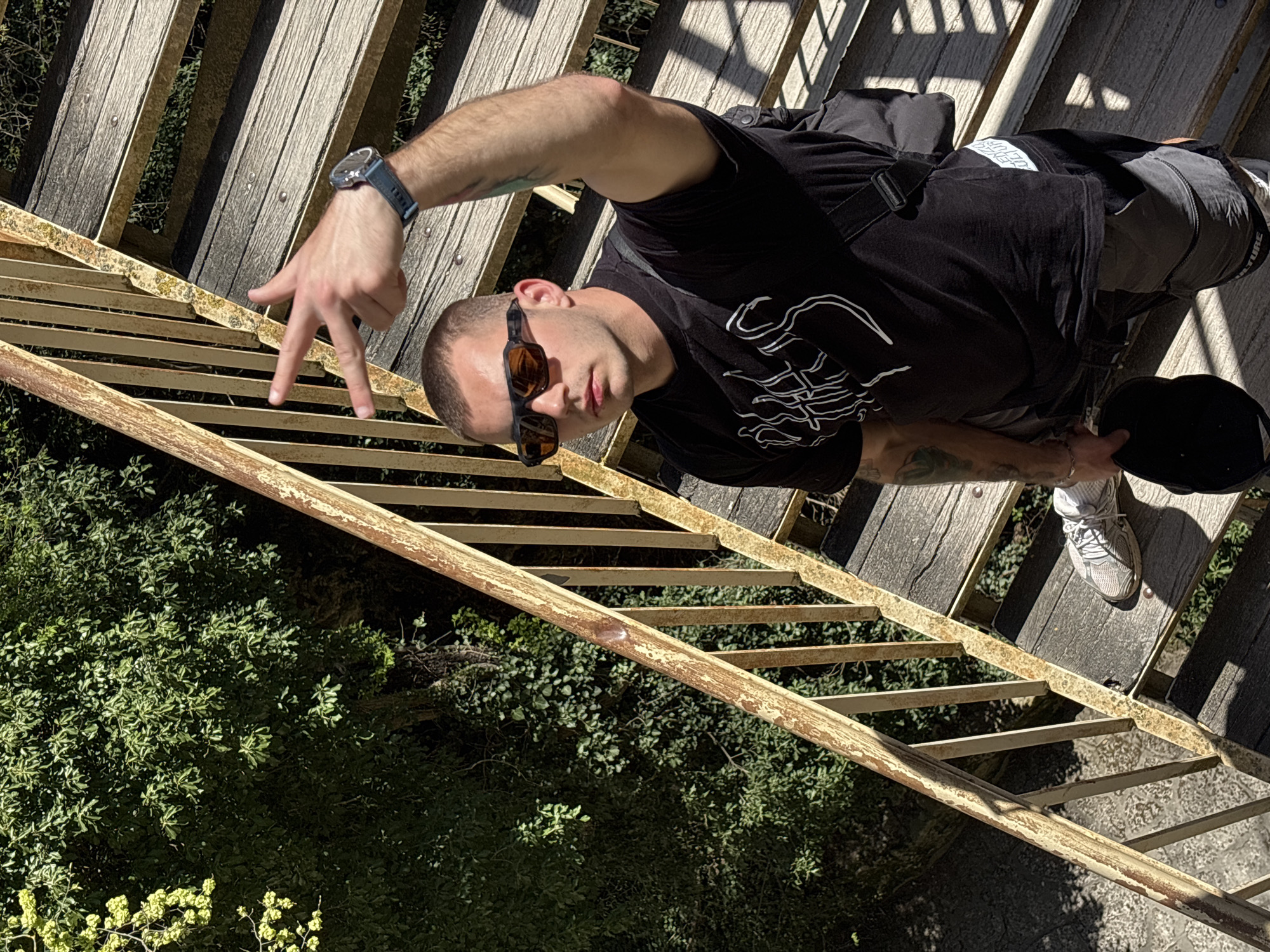 A man stands on a ladder, leaning towards the camera. He is wearing sunglasses, a black t-shirt with a white graphic, grey pants, and white sneakers. He has a watch on his left wrist. He is holding a black object in his right hand. His left hand is extended towards the ladder. The ladder is old and rusty, with wooden steps. Below the ladder is dense green vegetation. The location is Unknown, Unknown.