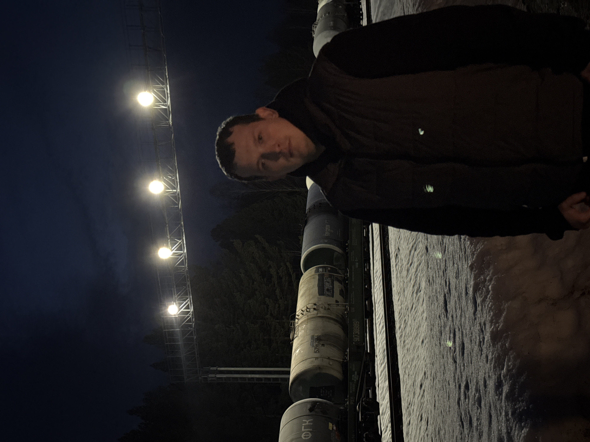 The image displays a nighttime scene at a railway line in Kemul, Russia, during winter conditions. In the foreground, a male individual stands, facing directly towards the camera with a neutral expression. He wears a dark long-sleeved shirt or hoodie beneath a dark brown or black puffer vest. The ground surrounding him and between the railway tracks is covered in a substantial layer of textured snow. Behind the individual, two parallel railway tracks are visible, upon which a train composed of multiple cylindrical tanker cars is stationary. These tanker cars are predominantly light-colored, with some displaying Cyrillic markings such as "ФГК" and numerical identifiers including "91738989" and "15396266". Beyond the tracks and train, a dense line of dark evergreen trees forms the horizon. Overhead, a metallic gantry structure spans the railway, supporting a series of bright, circular lights that illuminate the immediate area against a dark night sky with subtle cloud cover. No interactions between individuals or objects are depicted.