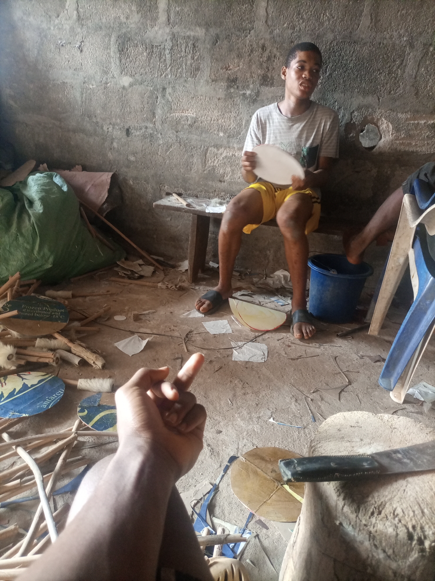 The image depicts an indoor scene in what appears to be a workshop or storage area. The walls are made of rough, unplastered concrete blocks. The floor is also concrete, strewn with various materials including wood scraps, paper, and what look like parts of unfinished drums or similar circular objects.
The primary subjects are two young men. One is seated on a low wooden bench, holding a white circular object, possibly a drum head, and looking towards the camera. He is wearing a striped t-shirt and yellow shorts. His posture suggests he might be in the process of working on the object. Another person is partially visible in the lower right corner, with their legs and feet visible. Their right foot is resting on the rim of a blue bucket. There are also many wooden sticks and cylindrical pieces scattered on the floor, along with several round objects with blue and yellow patterns on them, suggesting a craft or manufacturing environment. A green tarp-covered object is in the background, to the left. A sharp tool, possibly a chisel, is resting on a wooden surface in the foreground.
The overall impression is one of industrious activity in a somewhat cluttered and unfinished setting. The lighting suggests it is daytime, but there are no clear indicators of the specific time of day or weather outside. The expressions of the subjects are neutral or focused on the task at hand.
Visible text is present on some of the circular objects on the floor. One clearly reads "Frozen Product" and "stored below 0°C". Another has partial text that appears to include "antarcti".