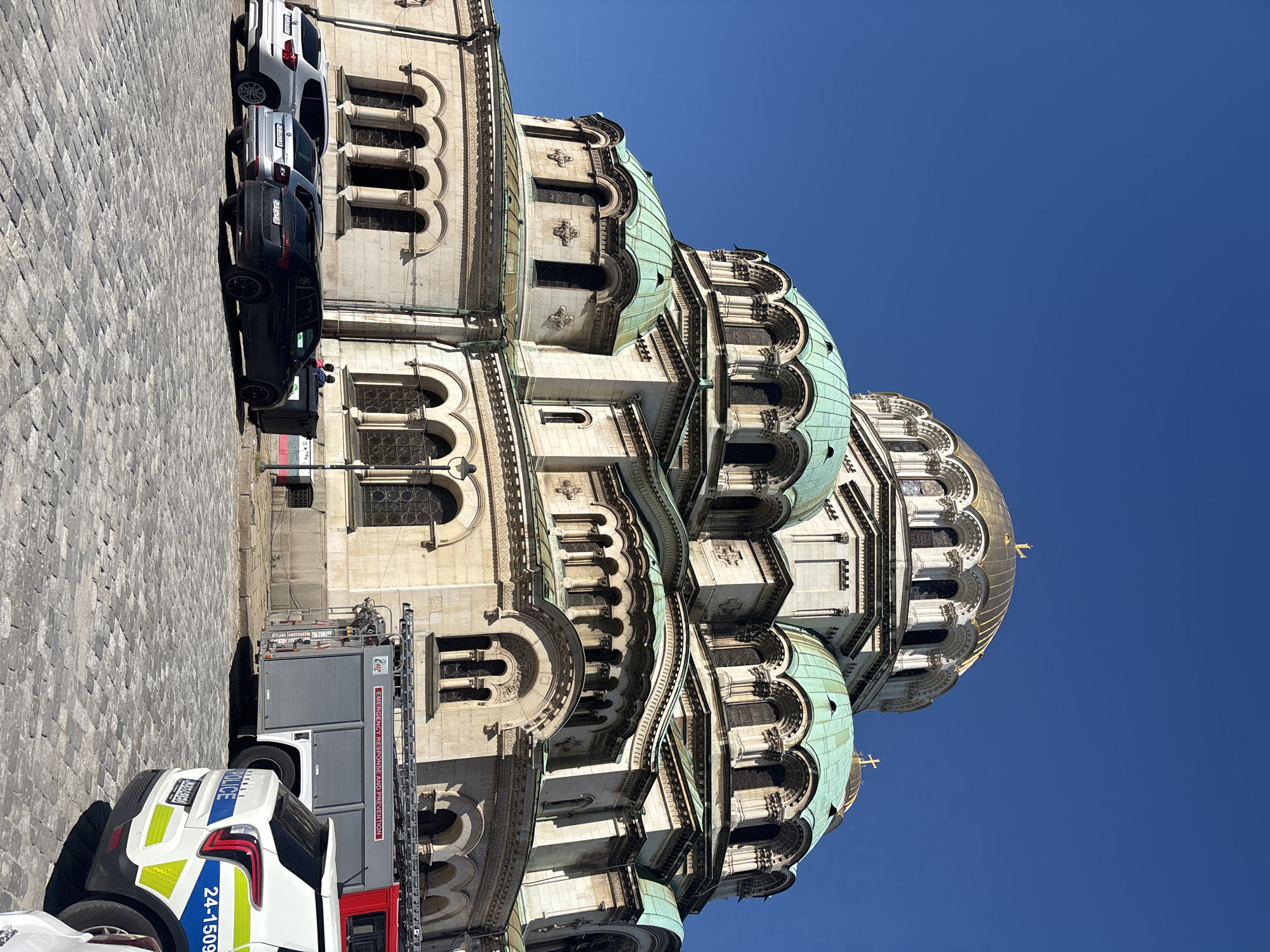 The image shows the Alexander Nevsky Cathedral in Sofia, Bulgaria. The cathedral is a large, ornate building with multiple domes, some of which are covered in green copper. Several vehicles are parked in front of the cathedral, including a police car and a fire truck with "EMERGENCY RESPONSE AND PREVENTION" printed on the side. The sky is clear and blue.