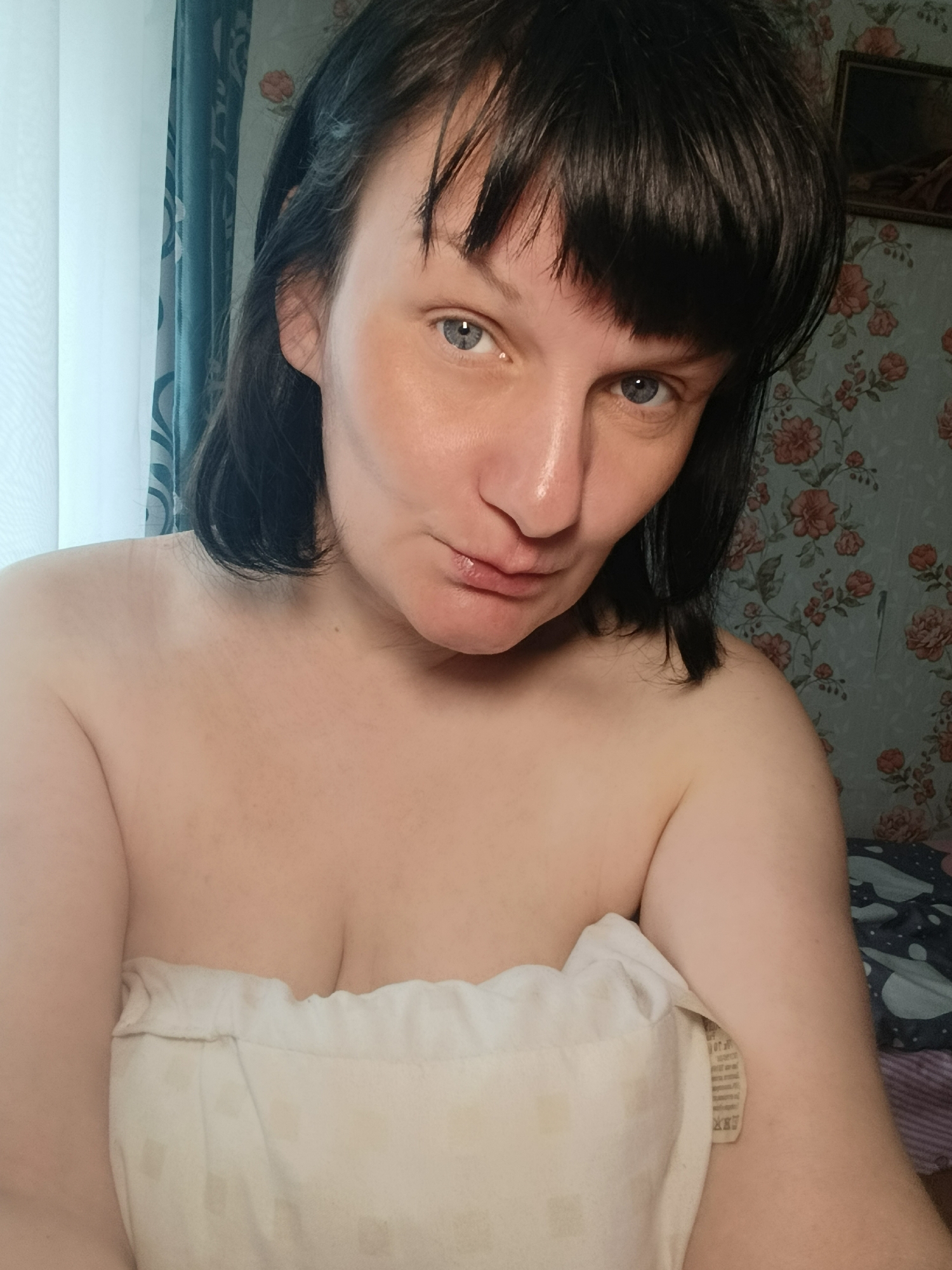 An indoor, close-up selfie captures an adult woman looking directly at the camera with blue-grey eyes and a subtle, playful pout. She has dark, short-to-medium length hair with bangs. Her shoulders and upper chest are bare, and she is wrapped in a plain white towel or blanket. Her skin shows a slight reddish flush on her cheeks and forehead, and her lips appear glossy.
The background on the right features floral wallpaper with a pattern of pink and orange roses on a light blue-green base, typical of a domestic setting in Фоки, Russia. In the upper right corner, a small, framed picture is visible on the wall. To the left, light sheer white curtains and darker blue patterned curtains indicate a window, suggesting natural light is present. A white tag with unreadable text is visible, sticking out from the side of the white towel or blanket. The overall lighting suggests it is daytime.