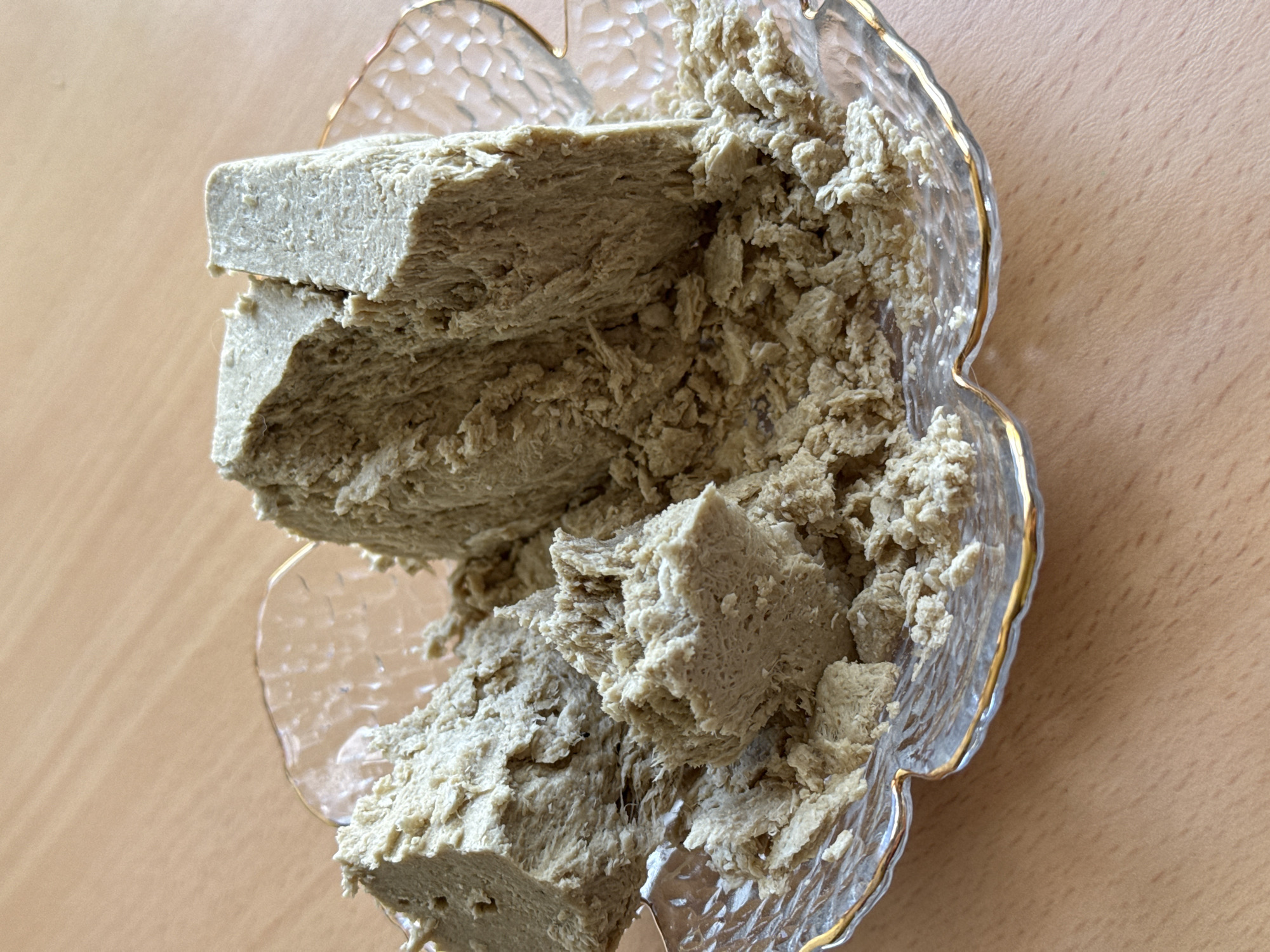 A close-up shot displays a bowl of halva on a wooden table. The bowl is clear, with a gold-painted rim and a flower-shaped edge. The halva is broken into irregular chunks and crumbled pieces. The background is a plain, light-colored wooden surface. The image was likely taken indoors under ambient lighting. There is no information to verify the location in Chaikovsky, Russia.