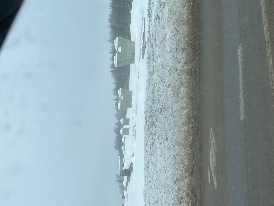 The scene depicts a winter landscape under an overcast sky, observed from within a vehicle, possibly in the city of Borisovo, Russia. The foreground shows a dark, wet asphalt road with a partial white line and a faded yellow marking, resembling a "7" or "Z," on its surface. Immediately adjacent to the road is a high, dirty snow bank composed of packed, brown-tinged snow.
Beyond the snow bank, the midground is dominated by a snow-covered, open construction site. Scattered across this terrain are multiple large, light grey concrete structures, identified as potential bridge supports or pillars. These structures vary; some are individual, wide-based pillars tapering upwards with concave, saddle-shaped tops designed to receive horizontal beams. Others appear as pairs of these structures. To the far left, a long, low, light-colored building is visible partially obscured by snow, alongside what appears to be a yellow piece of heavy construction equipment. Several reddish-brown objects, resembling timber or pipes, are lying on the snow to the right.
The background consists of a dense line of leafless trees forming a dark silhouette against the uniformly light grey sky, suggesting a wooded area or forest. No people are visible, and no active construction work or interactions are apparent. The environment is characterized by heavy snow cover and cold, diffuse lighting conditions typical of winter.
