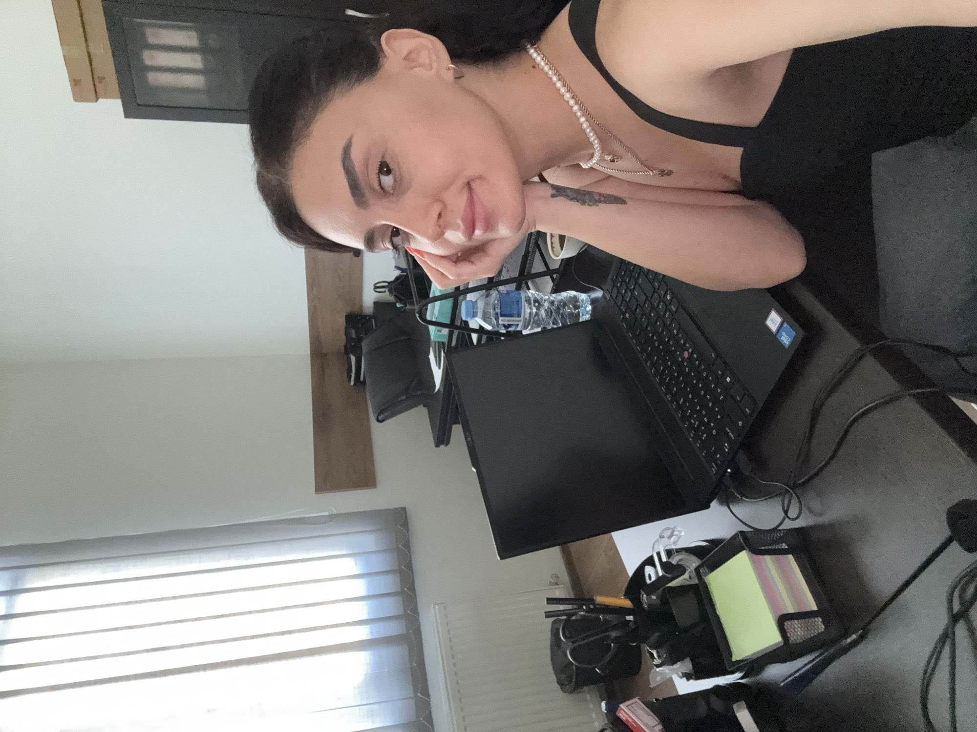A young woman with dark hair is sitting at a dark wooden desk in an office. She is looking at the camera with her hand resting on her chin, and she has a slight smile. She is wearing a black tank top and a pearl necklace with a pendant. On the desk in front of her is a black laptop, a bottle of water, a cup, and a holder with colorful sticky notes and pens. In the background, there is a radiator, a window with vertical blinds, a trash can, and a black cabinet. The overall setting appears to be a modern office space, possibly located in Tbilisi, Georgia, given the context.