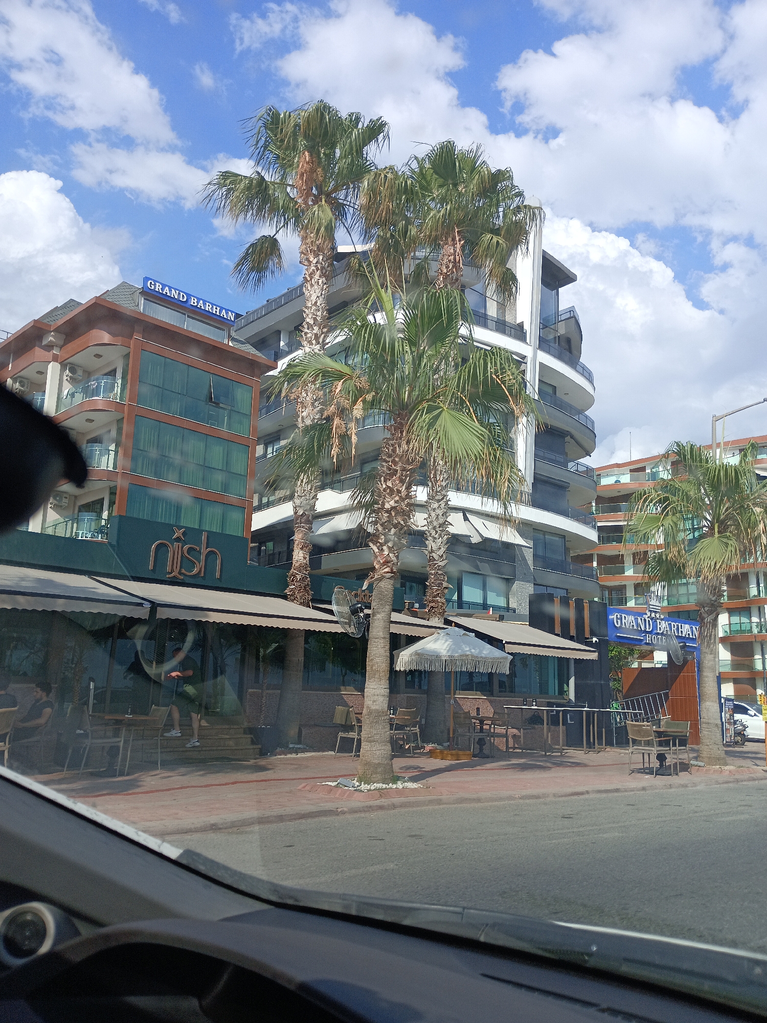 The image shows a street view in Alanya, Turkey, with a focus on a modern hotel and a restaurant. Dominating the scene are several tall palm trees. In the background, a multi-story hotel building with a curved facade and balconies is visible. A sign that reads "GRAND BARHAN HOTEL" is prominently displayed on the hotel. To the left of the hotel, another building with brown and green elements is present, featuring a sign with the word "nish" in a stylized font, likely indicating a restaurant or cafe. Outdoor seating with tables and chairs is set up on the sidewalk in front of the restaurant, some of which are covered by awnings. Several people are visible, including a person standing on steps leading into the "nish" establishment, and other individuals seated at tables. The street itself is paved, and there are sidewalks with some landscaping. The sky is blue with scattered white clouds. The perspective suggests the photo was taken from inside a vehicle, possibly looking through the windshield, as the dashboard is partially visible in the foreground.