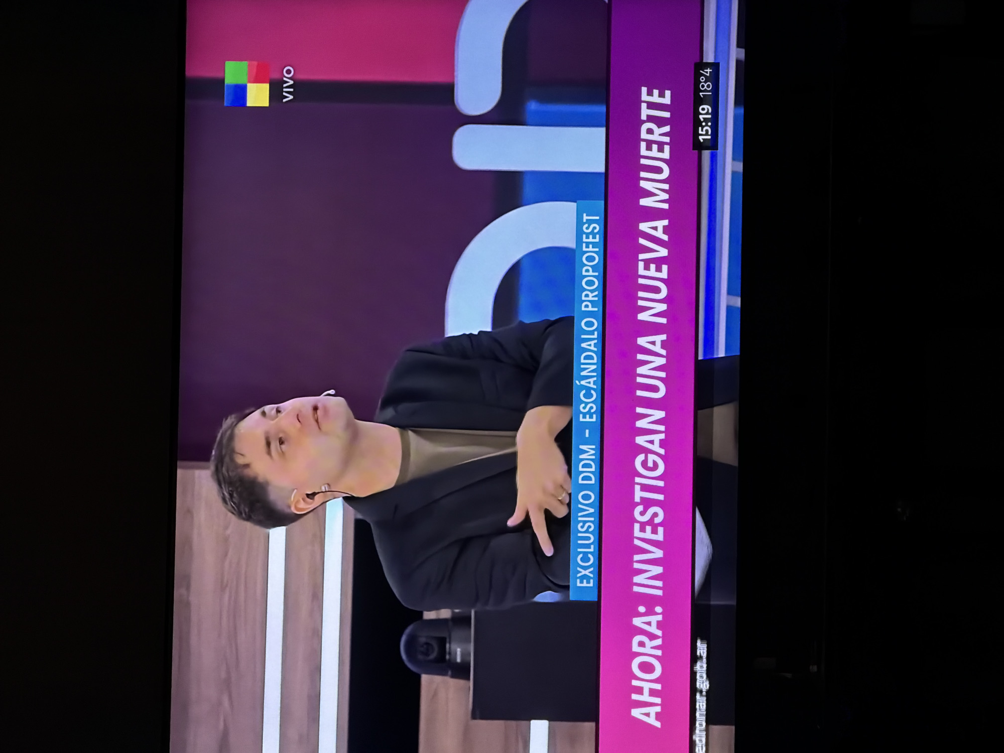 Here is a matter-of-fact description of the image:
The image shows a television screen displaying a news program. A man in a black suit stands on a stage. The background is a mix of maroon and light wood paneling with white vertical stripes. A camera is visible in the lower center. On the right side of the screen, there is a vertical purple bar with white text: "AHORA: INVESTIGAN UNA NUEVA MUERTE" and "EXCLUSIVO DDM - ESCÁNDALO PROPOFEST". At the top left, there is a logo reading "VIVO". In the top right corner, the time "15:19" and temperature "18°4" are displayed. At the bottom of the text bar, a URL is visible "edronar..gob.ar". The image appears to be a recording or photograph of a television broadcast.
