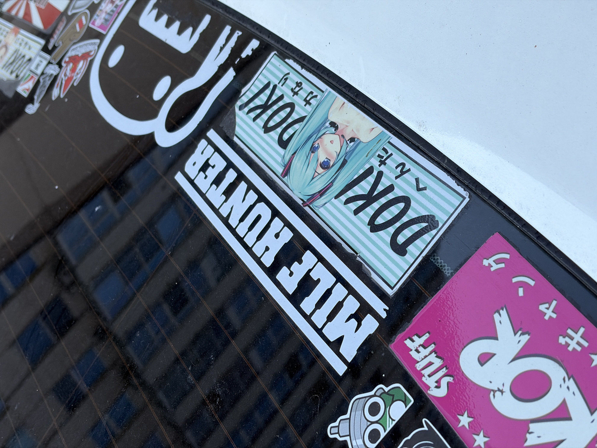 The image shows a close-up view of stickers on the rear window of a vehicle. The window has a grid pattern from defroster lines. The stickers include a large white graphic of a stylized black character with a wide mouth and large eyes, positioned on the right side of the frame. To the left of this character, there is a diagonal white sticker with the text "MILE HUNTER" in large, bold font. Below and to the left of "MILE HUNTER," there is a smaller sticker with a green and gray illustration of a gas mask-like figure. In the lower portion of the frame, a bright pink sticker features the text "STUFF" in white at the top and a large white stylized letter "R" with jagged edges. To the right of this pink sticker, a horizontal sticker displays a light blue anime character with long turquoise hair and blue eyes, positioned next to the text "DOKI" and some Japanese characters. The overall environment suggests an outdoor setting, likely a parking area, with the reflection of a building visible on the car window. There is no indication of the specific location within the city of Khimki, Russia.