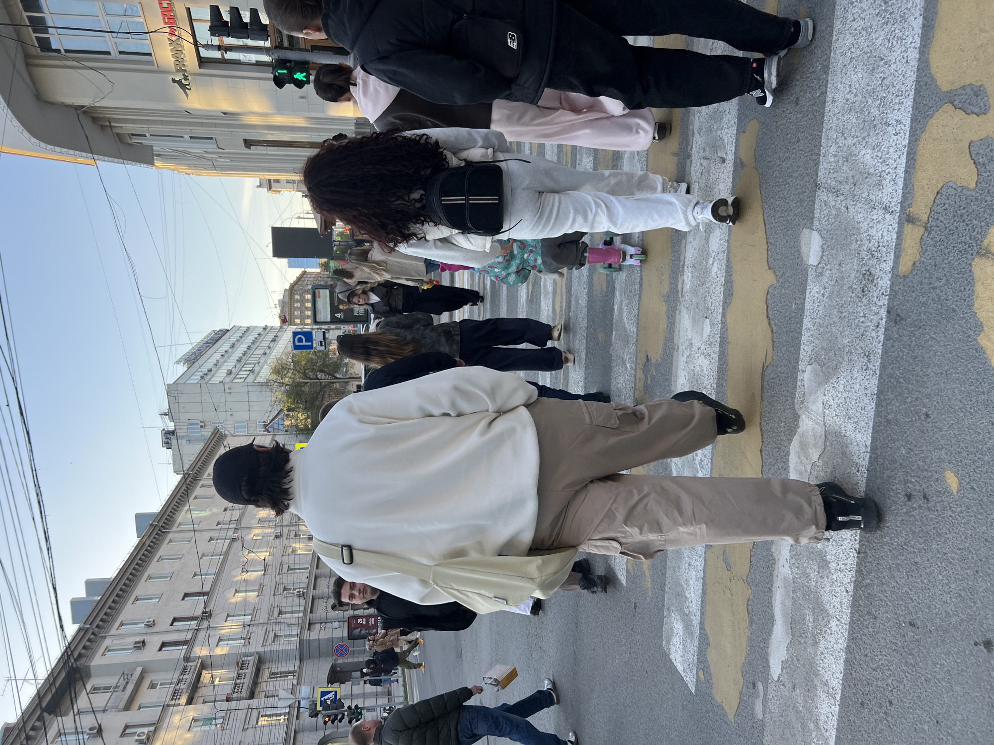 The image shows a pedestrian crosswalk scene in Novosibirsk, Russia. A group of people is crossing the street, moving from the bottom towards the top of the frame. The crosswalk has gray and yellow stripes. In the foreground, a person in a white sweater and khaki pants is crossing, with their back to the camera. To their right, a man in a green jacket and jeans is walking. Various other people are visible in the crosswalk, including a person with curly hair wearing white clothes. Buildings line the street on both sides, and traffic lights are visible in the top left and bottom left corners, showing a green light for pedestrians. Overhead wires are strung across the sky.