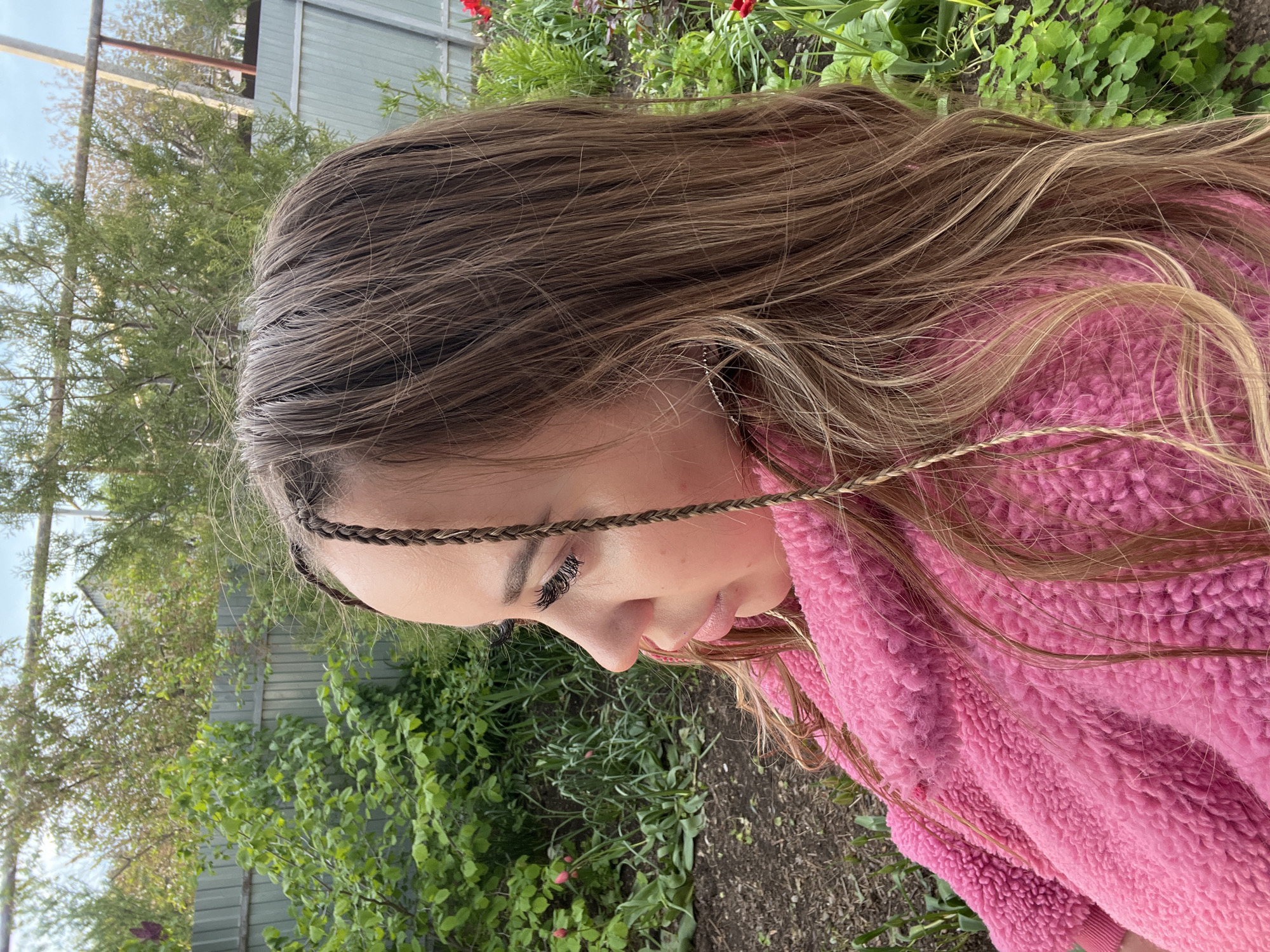 A young woman with long, light brown hair and a braid across her forehead is outdoors. She is wearing a pink fleece jacket and looking down. The background includes green foliage, a wooden fence, a tree, and part of a building. The location appears to be a garden or yard, and it is in Novopokrovskaya, Russia.
