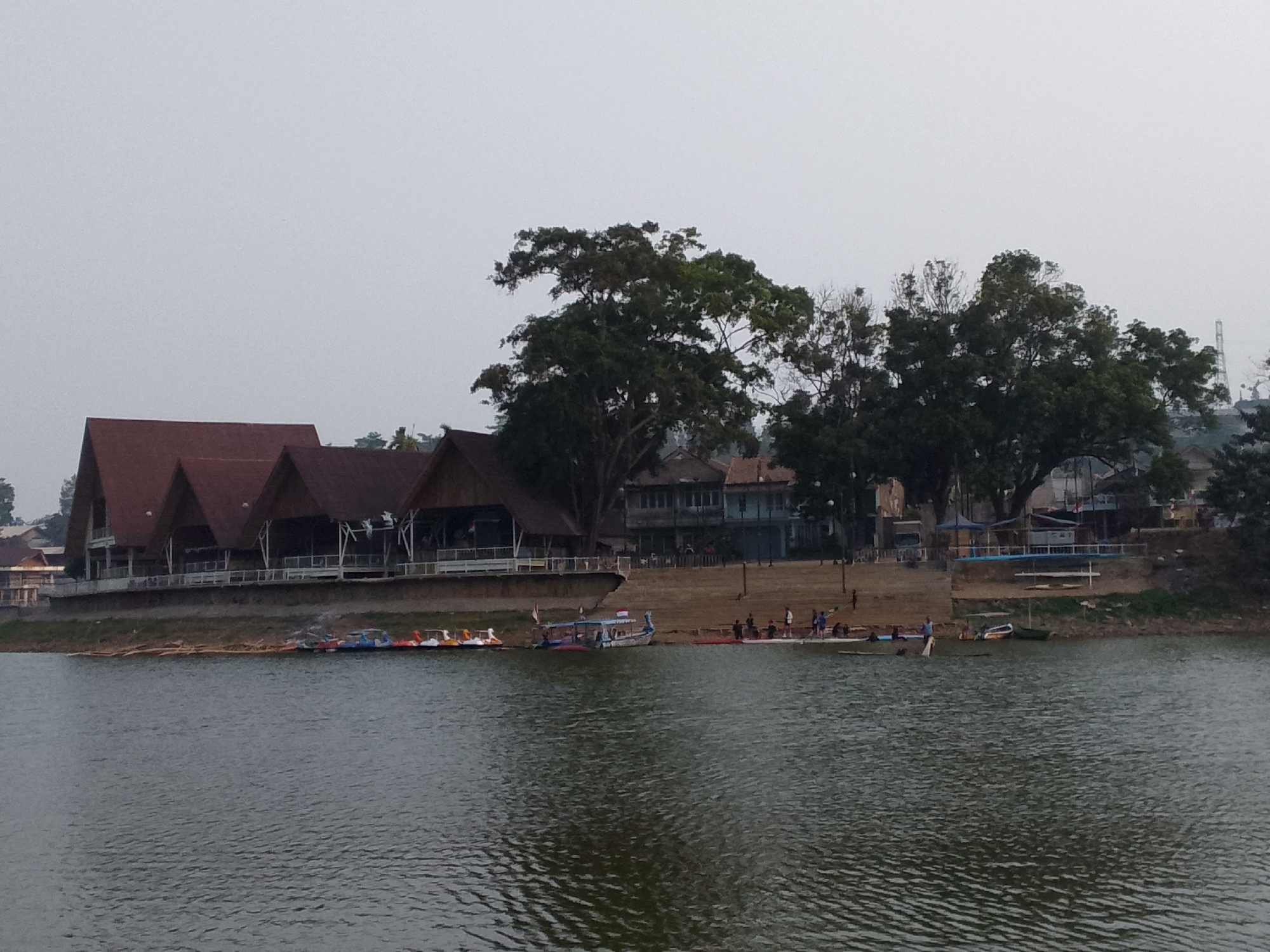 This image depicts a waterfront scene at the Situ Ciburuy lake in Padalarang, Indonesia, under an overcast sky. In the foreground, the dark, rippling surface of the lake occupies the lower half of the frame. Along the shoreline, several small boats are visible. These include at least five brightly colored paddle boats lined up on the left side, and a larger, more traditional boat with a blue and white canopy to the right of the paddle boats. Further down the shore, a green canoe is beached.
Behind the shoreline, a grassy bank slopes upwards towards a row of buildings and trees. The most prominent structures on the left are a series of interconnected buildings with distinctive, steeply pitched, reddish-brown roofs, likely part of a restaurant or resort. Further to the right, a light blue, multi-story building is visible, and behind it, several large, mature trees with dense green foliage dominate the middle ground. Scattered individuals are present along the shore, some standing near the boats, and others appearing to walk or gather. The overall impression is of a recreational area on a cloudy day.