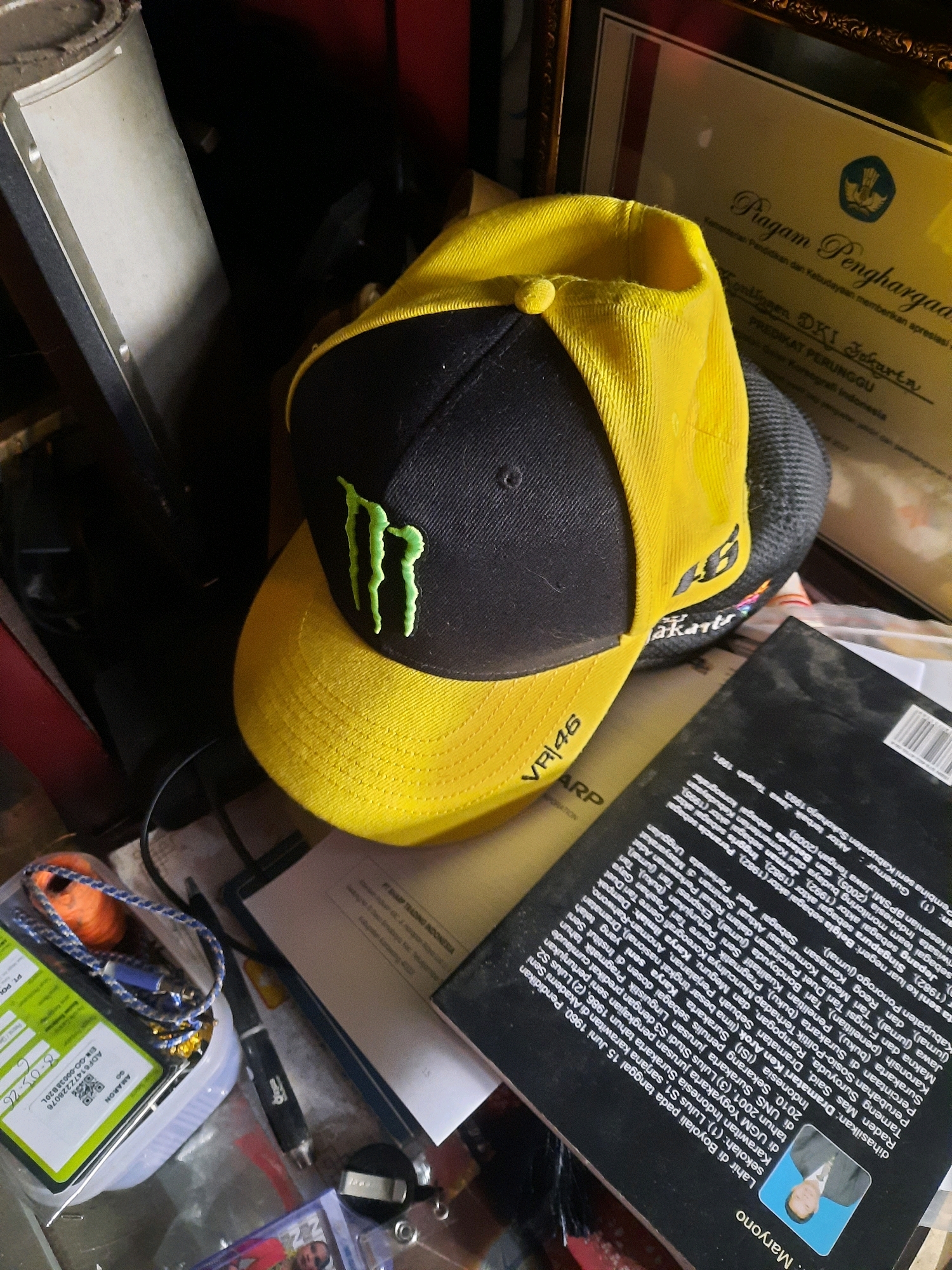 A yellow and black baseball cap with a Monster Energy logo is displayed on a surface cluttered with various objects. The cap is angled, with the yellow brim facing forward and the black crown tilted to the left. Behind the cap, a framed document with Indonesian text and the Garuda Pancasila emblem is visible. To the right of the cap, a black book with text in Indonesian is partially open, revealing a photograph of a man in a suit. In the foreground and to the left, a small plastic container holds a lanyard with orange and blue string, a blue USB drive, and other small items. A pen and a silver object are also on the surface. The environment appears to be indoors, likely a desk or shelf, with a red background element. The presence of the framed document and the Indonesian text suggests a location in Indonesia. Given the specific details of the framed award referencing "DKI Jakarta" and "Pemerintah Provinsi DKI Jakarta", it is highly probable that this scene is located within the city of East Jakarta, Indonesia.