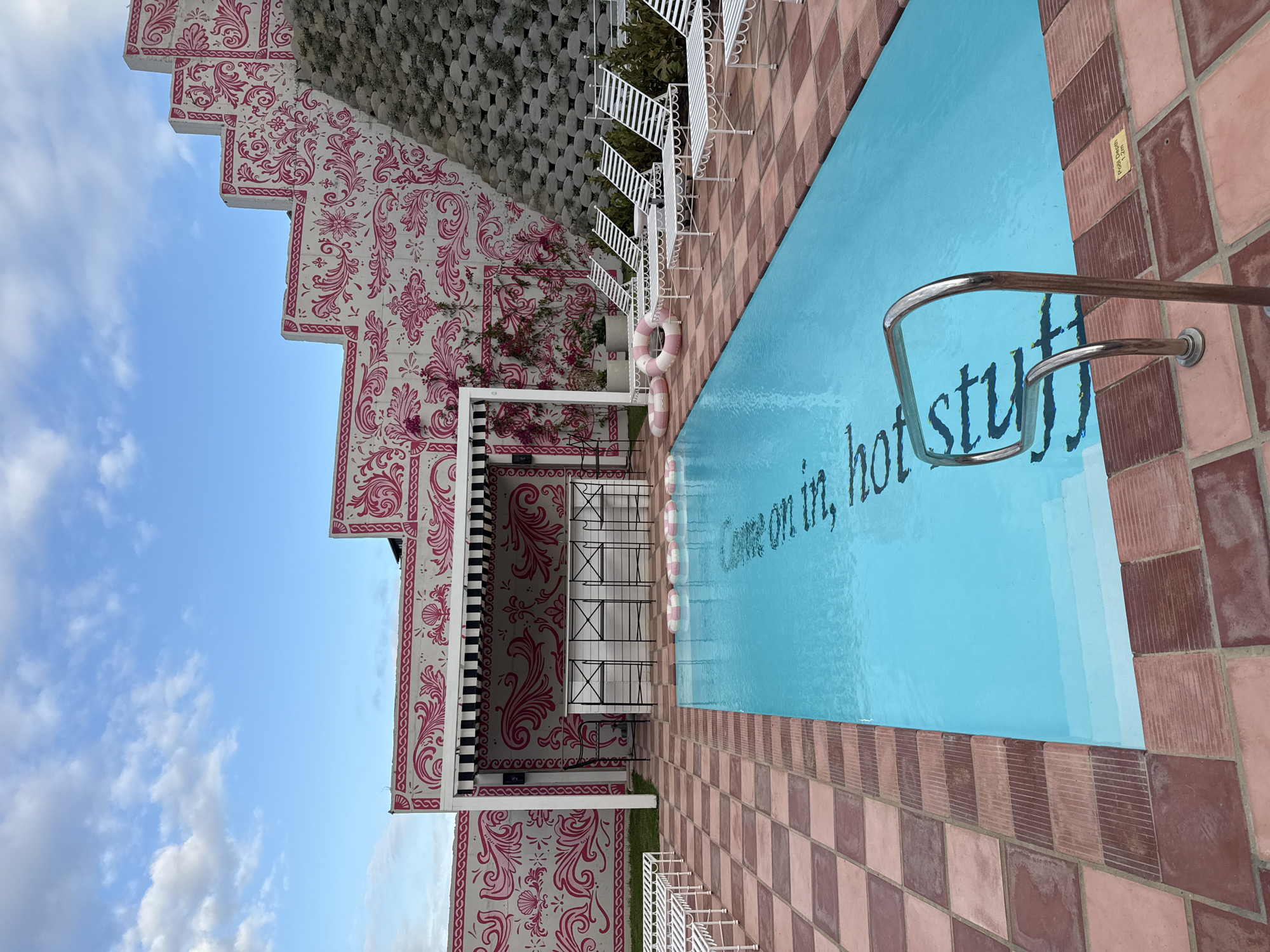The image depicts a swimming pool at The Silo Hotel in Cape Town, South Africa. The pool is rectangular with light blue water, and the words "Come on in, hot stuff" are written on the bottom. The pool is surrounded by checkered tile and a low metal railing. A pink and white patterned building is adjacent to the pool area, and there are white chairs visible. The sky is blue with some clouds.