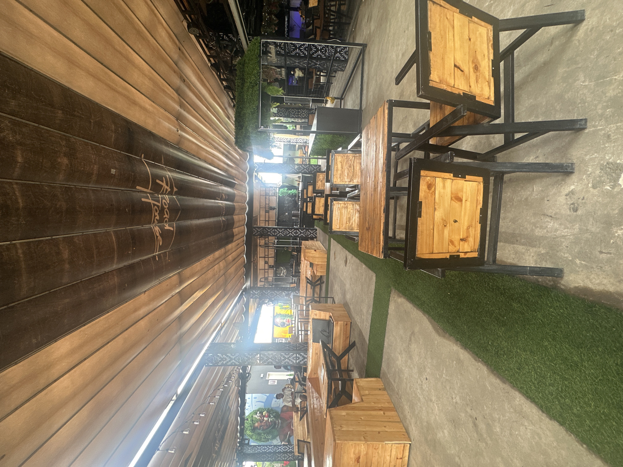 This is a medium-shot, eye-level, outdoor photograph of a cafe or restaurant seating area in Howayi, Nigeria. The scene is dominated by wooden tables and chairs with black metal frames, arranged on concrete and artificial turf flooring. A long, slatted wooden awning covers a portion of the seating area, with subtle lighting visible beneath it. The awning has dark brown wooden slats at the bottom and lighter wood slats above, with text that appears to read "Road House" in a stylized font. In the background, there is more seating, some covered by a more permanent structure with decorative metal panels. A large mural or advertisement is visible on a wall in the distance, featuring colorful imagery. The setting appears to be an establishment that is either open or preparing to open, with tables set and ready for customers. The time of day is difficult to ascertain precisely, but the lighting suggests it could be late afternoon or early evening, possibly after sunset given the artificial lighting. The weather appears to be clear. There are no people prominently featured in the foreground, but several individuals are visible in the background, appearing to be staff or patrons. The overall impression is one of a modern, casual dining space with an industrial-chic aesthetic.