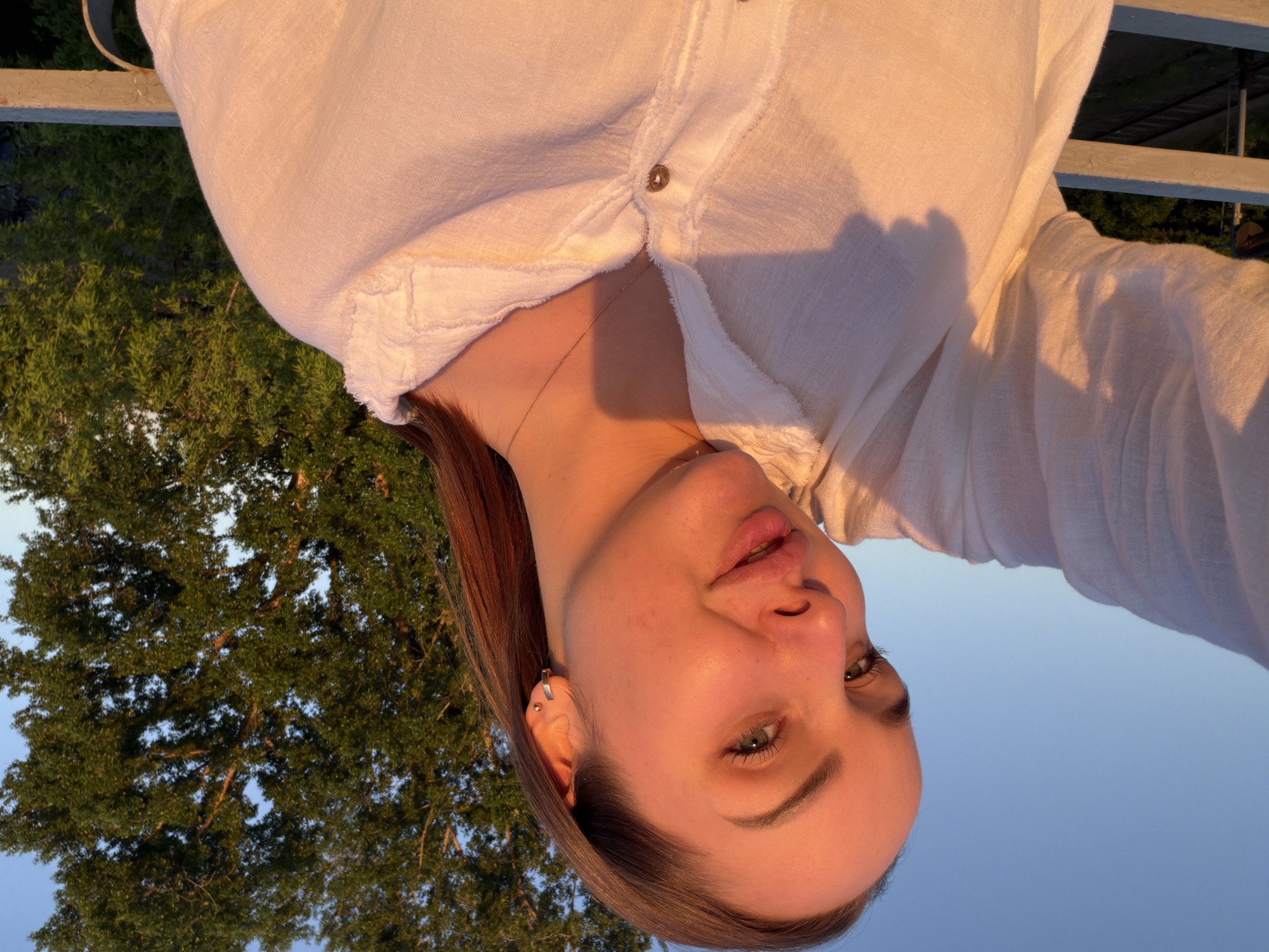A person with light skin and brown hair wearing a white button-down shirt is facing the camera. The background shows trees and a blue sky, suggesting an outdoor setting possibly in the city of Chaikovsky, Russia, given the request. A metal railing can be seen partially at the top of the frame. The image is rotated.
