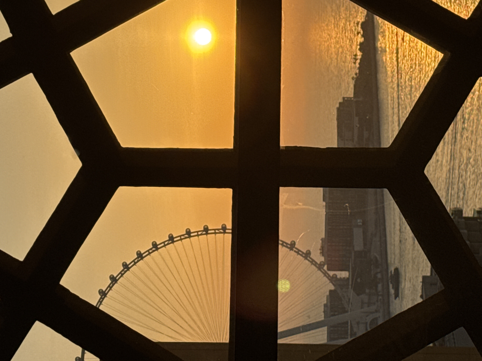 The image captures a sunset in Dubai, United Arab Emirates, viewed through a geometric window frame. In the upper portion of the frame, the iconic Ain Dubai observation wheel is partially visible against a warm, orange sky, with the setting sun shining as a bright orb. The lower portion of the frame shows a body of water reflecting the sunset's golden hues, with buildings and a small boat in the distance. The overall scene evokes a serene and picturesque end to the day in the bustling city.