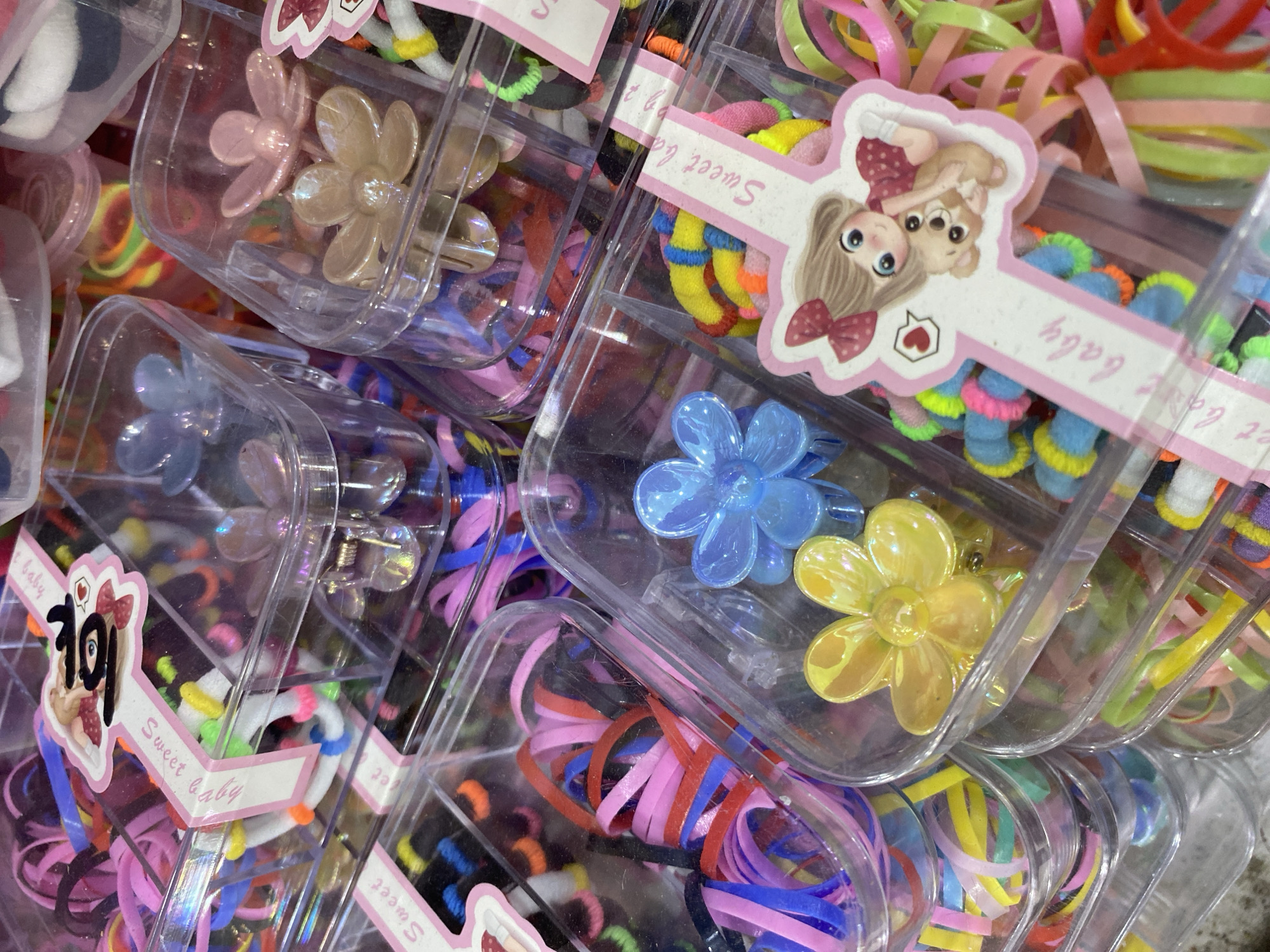 In the city of Lamteumen Barat, Indonesia, a close-up, overhead shot captures a retail display of hair accessories, predominantly hair ties and flower-shaped clips. The items are neatly organized in clear plastic containers, stacked on shelves or in bins, suggesting a store environment. The hair ties are a vibrant mix of colors, including pink, red, blue, yellow, green, and black, bundled together in various containers. Some containers hold only hair ties, while others feature decorative elements like flower-shaped hair clips in translucent blue, yellow, and pink. One prominent container displays a pink sticker with a cartoon illustration of a girl hugging a teddy bear and the text "Sweet baby" in English, indicating a product marketed towards children. The lighting is bright, illuminating the colorful accessories and the clear plastic packaging, creating a visually appealing display. The scene is static, with no people or discernible activity other than the display of merchandise. The background is out of focus, with hints of more products and retail shelving.