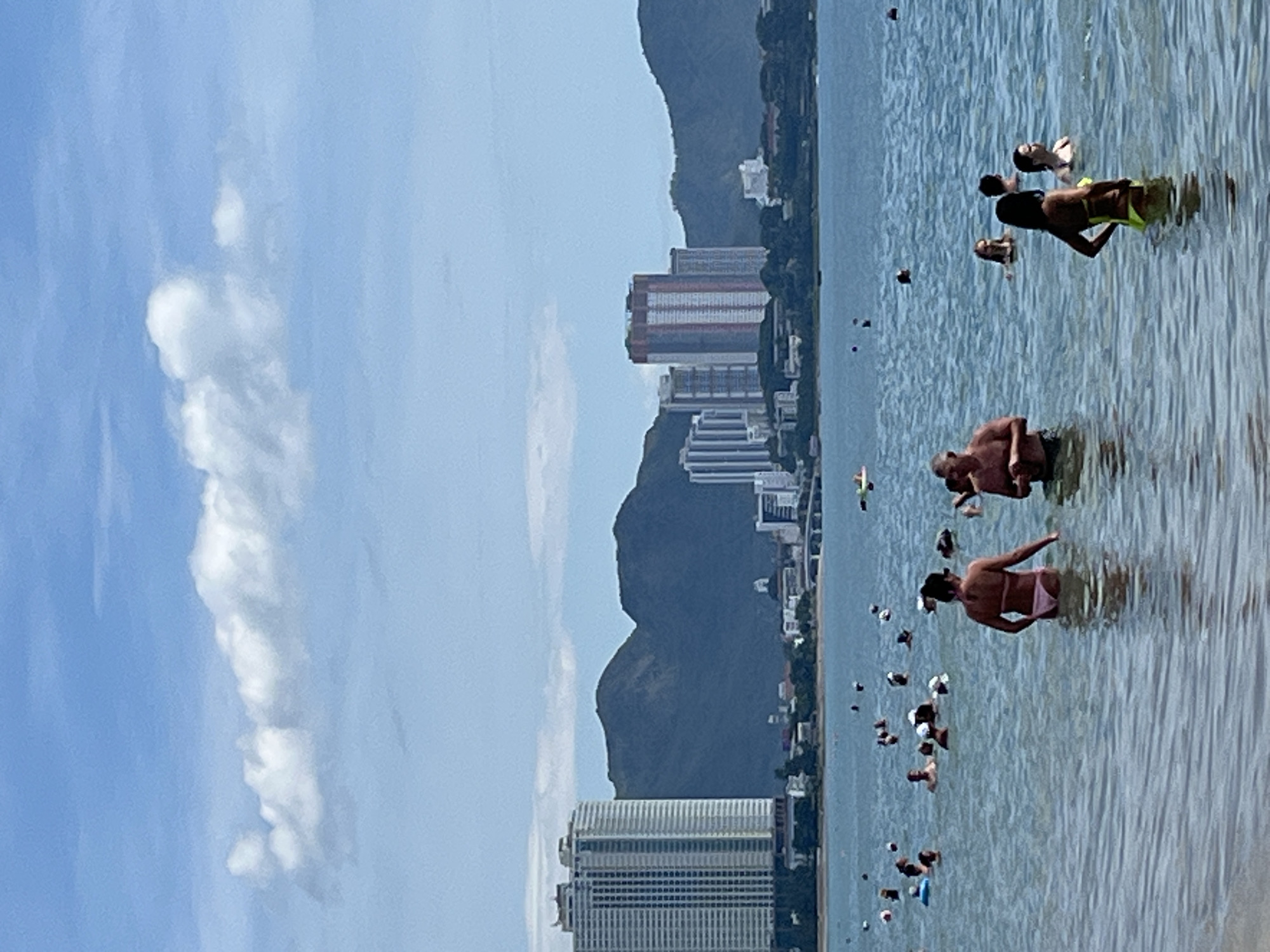 The image depicts a crowded beach scene in Nha Trang, Vietnam. In the foreground, several people are wading in the clear blue water. A woman in a pink bikini is facing away from the camera, and a man in swim trunks is next to her. Further out, more individuals are swimming and enjoying the water. In the background, a city skyline with tall buildings and mountains can be seen. The sky is partly cloudy, with white clouds against a bright blue backdrop. The overall impression is a sunny day at a popular tourist destination.