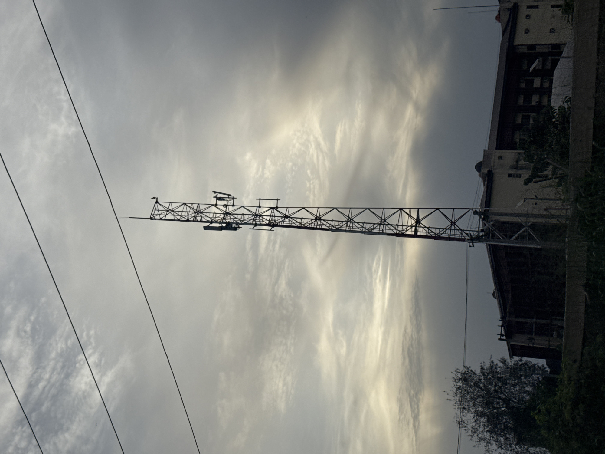 A low-angle, overcast shot shows a horizontal lattice boom crane pointing left across the frame. Thin, dark power lines cross diagonally in the foreground on the left. In the background is a building with multiple floors on the right, partially obscured by vegetation. The sky is overcast with striated clouds. The location is Obinze, Nigeria.