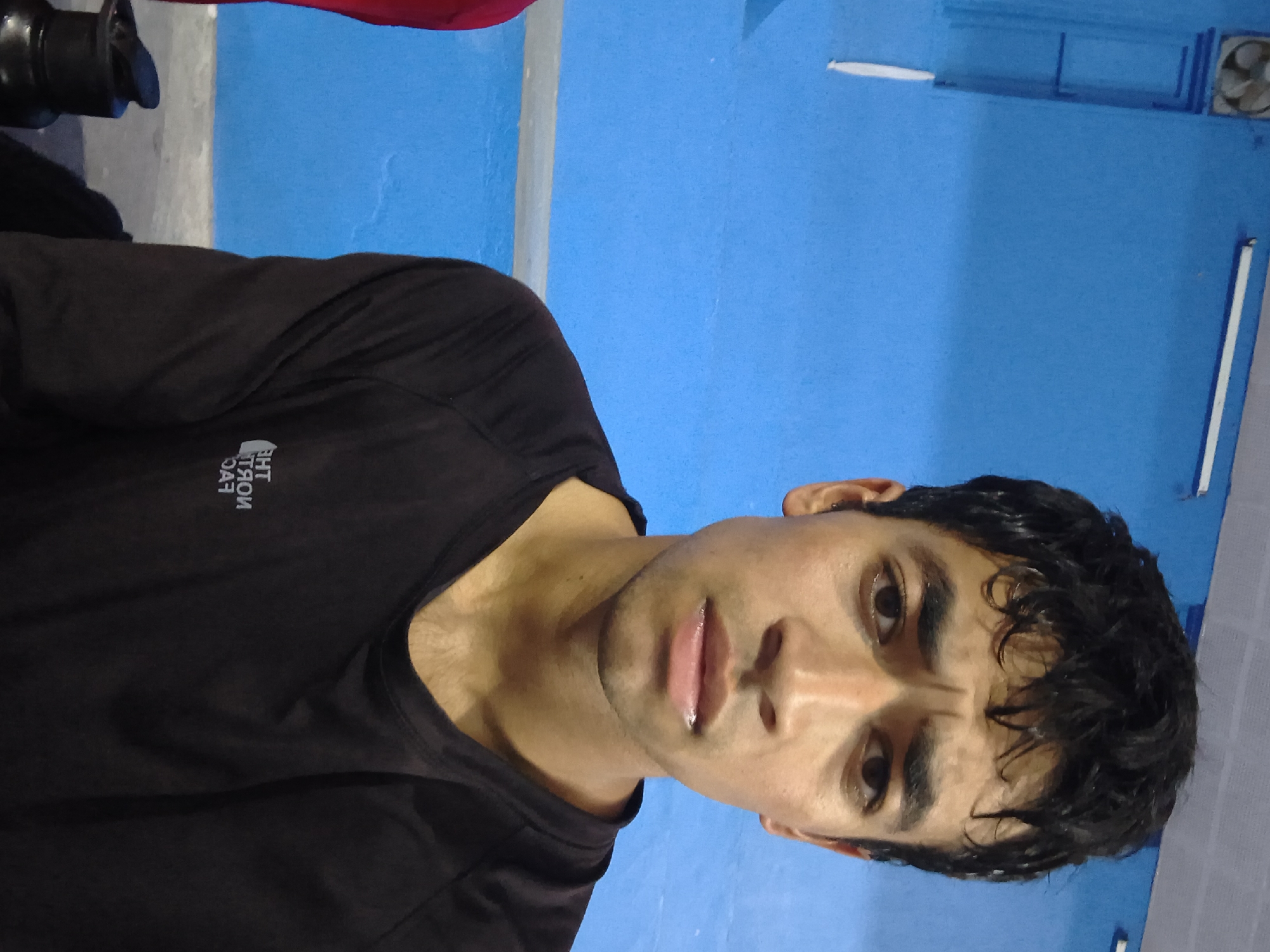 A young South Asian male is captured in a selfie-style photograph against a bright blue wall. He is wearing a dark long-sleeved shirt with a faint white logo on the left chest, which appears to be "THE NORTH FACE" reversed. The male's eyebrows are furrowed, suggesting a concerned or questioning expression. His hair is dark and slightly messy. In the background, on the upper left, a circular fan is visible, mounted on the blue wall. A horizontal grey bar runs across the wall, dividing the upper and lower sections. The bottom part of the image shows the male's torso and a portion of a black water bottle with a black lid on the left side. The scene appears to be indoors, possibly in a sports facility or gymnasium in Dharamshala, India, given the blue walls often found in such establishments and the presence of the fan.