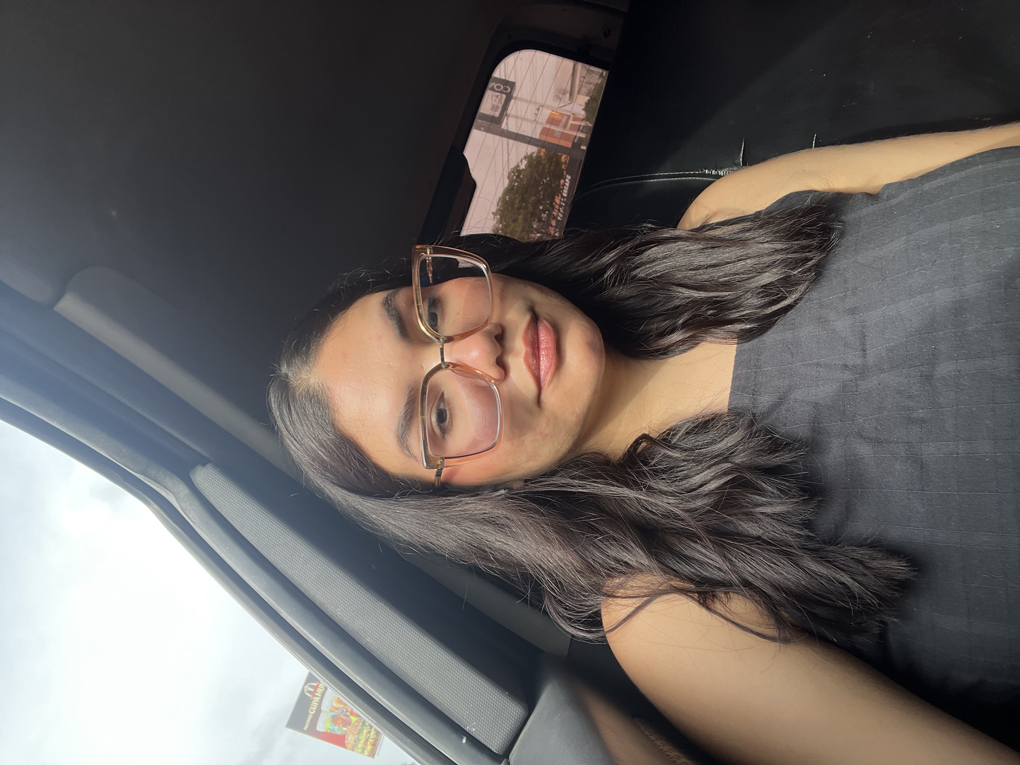 A medium shot shows a young woman with long, dark, wavy hair wearing large, square, beige-framed glasses and a dark, sleeveless top. She is inside a vehicle, with the interior of the car visible in the foreground and background. The woman is looking directly at the camera, with her head slightly tilted. Through the car window, a billboard advertising McDonald's and trees are visible, suggesting an outdoor, urban setting in Porlamar, Venezuela. The lighting is natural, indicating it is daytime, and the sky appears overcast.
