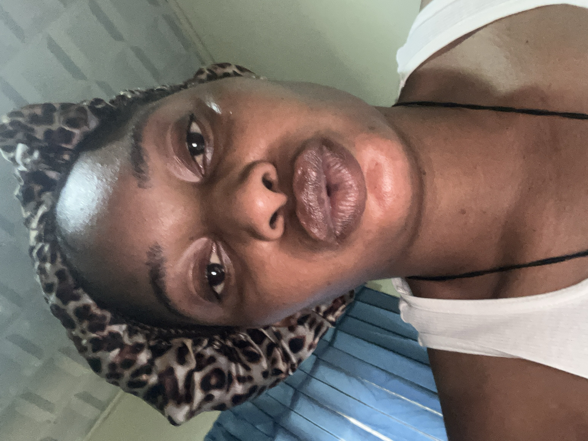 In the city of Jos, Nigeria, a person is captured in a close-up, front-facing photograph. They are wearing a white tank top and a leopard-print bonnet, styled with braided hair peeking out. The background is a light green wall with a ceiling that has textured panels. To the left, blue-green blinds are partially visible, suggesting an indoor setting. The person's face is centrally positioned, with their lips pursed in a kiss. Their eyes are open, and their skin appears moist, possibly from sweat or skincare products. A dark string, likely a necklace, is visible against their chest. The lighting suggests it may be daytime, with some ambient light reflecting off the person's forehead and cheeks.