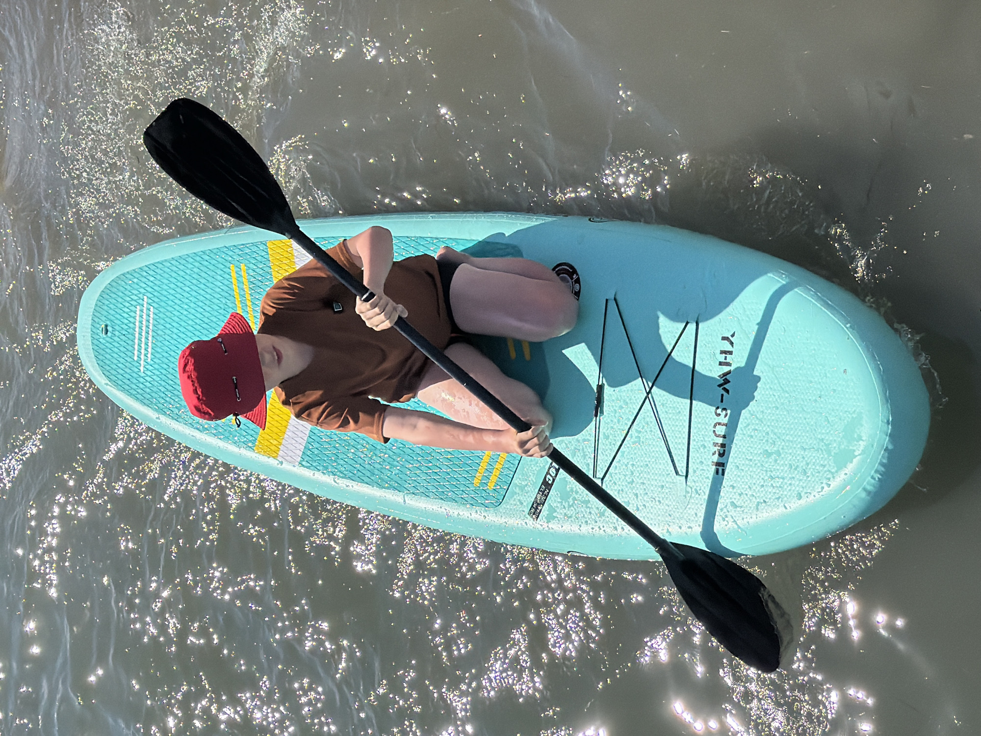 An overhead shot captures a person kneeling on a light blue paddleboard in rippling water. The person wears a brown t-shirt, dark shorts, and a red cap. Their legs are bent, with their right leg forward and their left leg tucked underneath. They hold a black paddle with both hands, positioned across the paddleboard. The paddle's blade is in the water on the right side of the frame. The paddleboard has yellow and white stripes on its surface. Text is visible on the paddleboard, reading "YHW-SURF". The water reflects the sunlight, creating a sparkling effect. The scene suggests a calm day outdoors, likely during daylight hours.