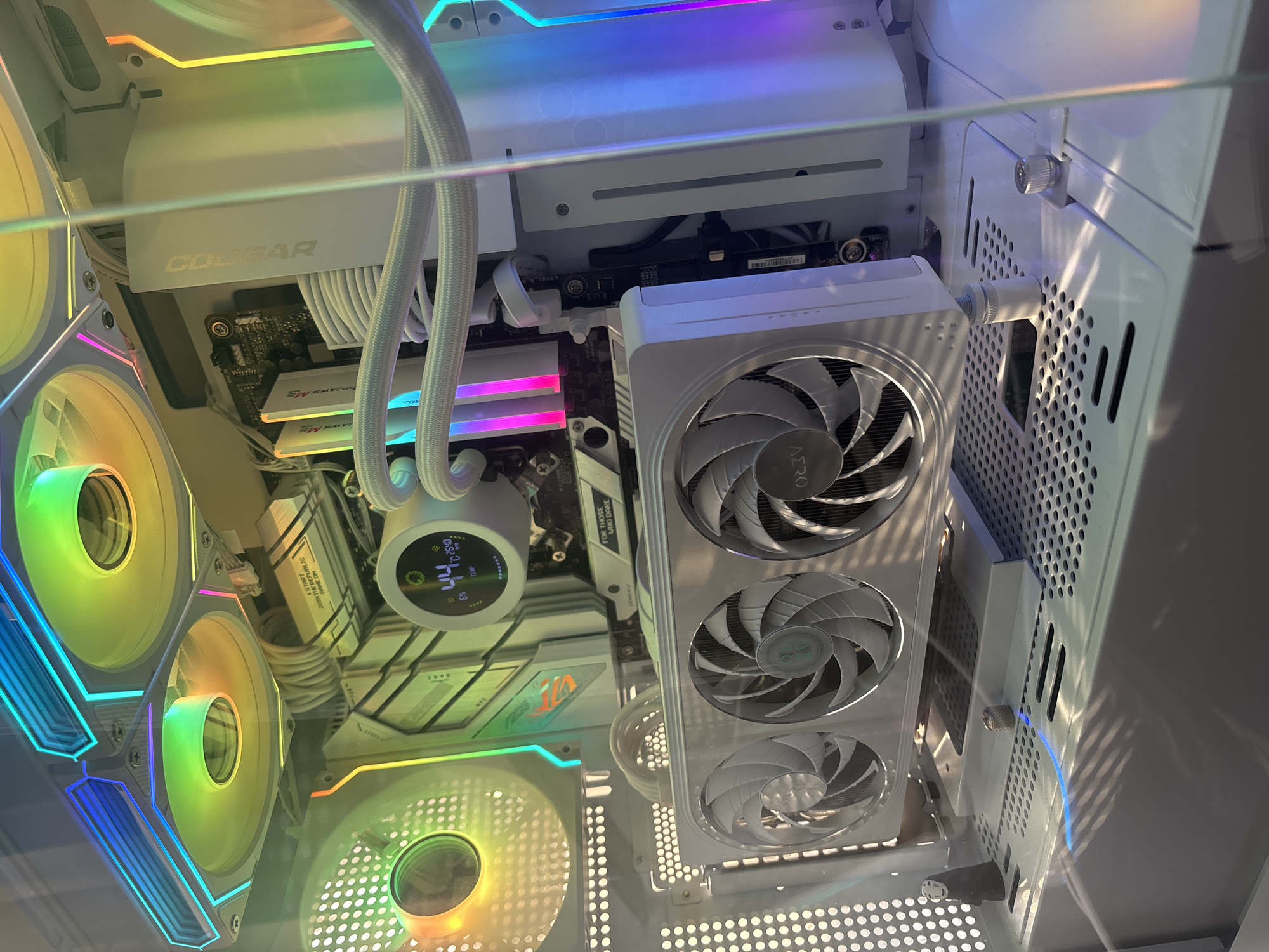 This is a close-up, high-angle shot of the interior of a white computer case. The primary subjects are the internal computer components, illuminated by RGB lighting that casts vibrant colors across the scene. Visible components include multiple computer fans with RGB lighting, a CPU cooler with a circular display showing "6% 44° TEMP", white cable management, RAM sticks with pink RGB lighting, and a large white component that appears to be a graphics card. Text visible on components includes "COUGAR", "THE REPUBLIC", "JOIN THE REPUBLIC", "GAME ON", "FOR THOSE WHO DARE", and "AERO". The scene is a controlled environment, likely a desk or workbench, with no visible indication of time of day, weather, or specific location beyond it being a computer build. The overall impression is one of a modern, high-performance gaming or workstation PC.