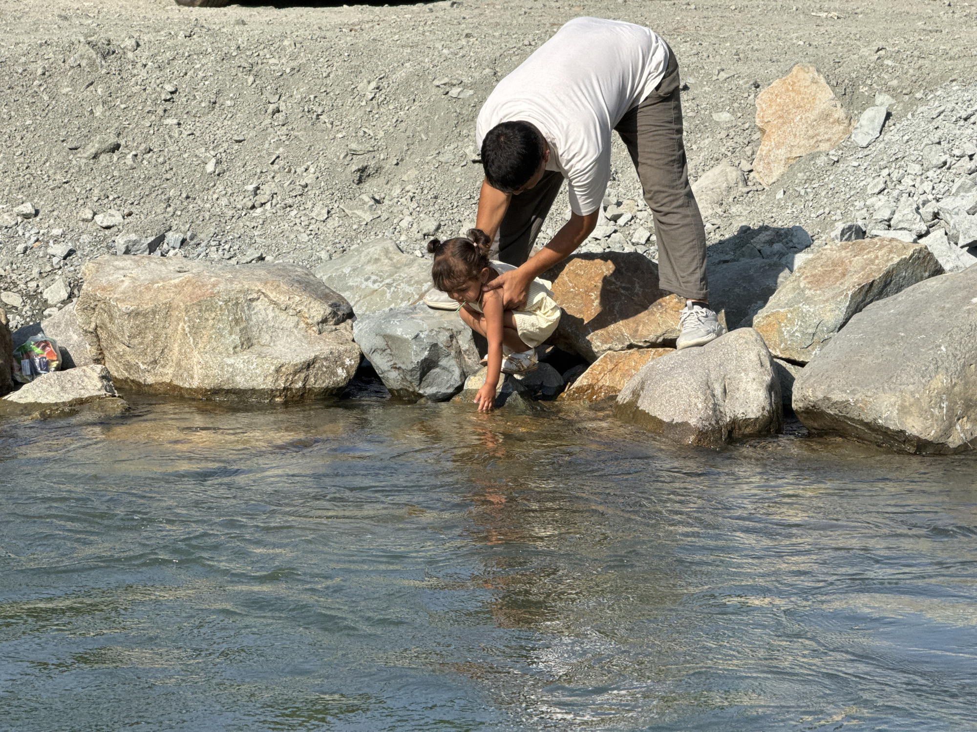In Azgilli, Azerbaijan, a man in a white t-shirt and dark pants is bending over to assist a young child in a light-colored dress. The child is crouching near the edge of a body of water, reaching a hand towards it. They are surrounded by large rocks and a gravelly embankment. The water is clear and appears to be flowing. The sun is shining, casting shadows on the ground.