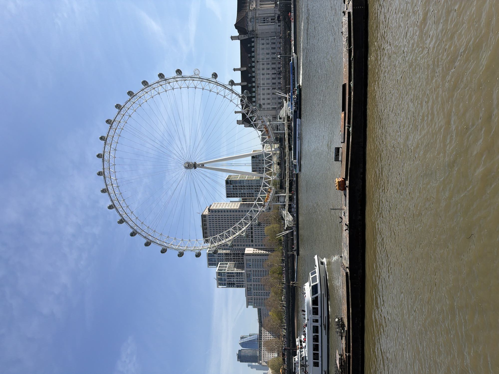 The image captures the London Eye, a large Ferris wheel, against a backdrop of buildings under a blue sky with wispy clouds. A river runs along the side of the image with a boat on it. The buildings behind the London Eye vary in height and architectural style, from modern glass structures to older, more traditional buildings. The river has a murky brown color. A metal structure runs parallel with the river and appears to have a boat docked next to it.