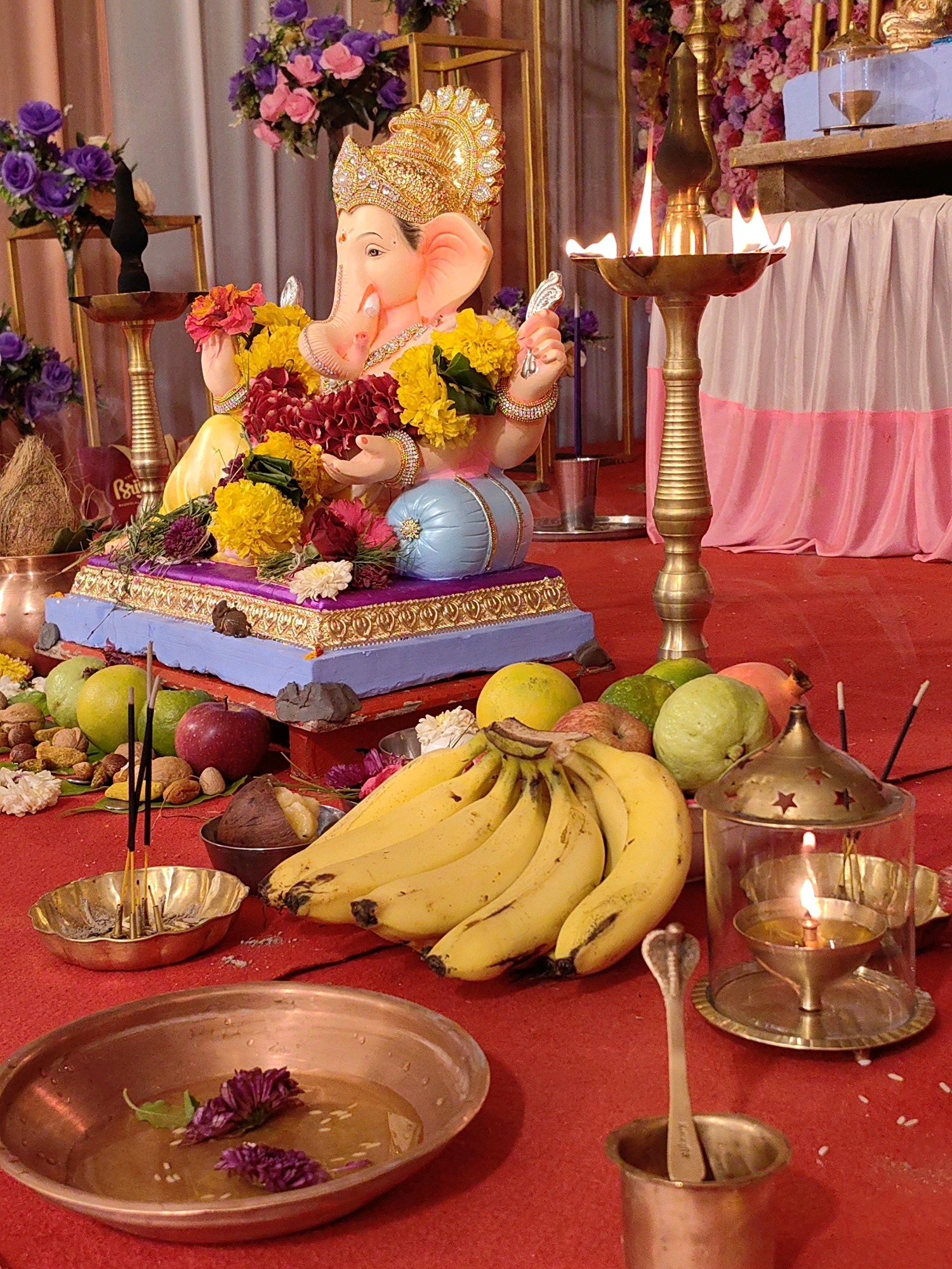 An indoor scene captures a religious offering dedicated to Lord Ganesha. A brightly colored idol of Ganesha, adorned with flowers and a golden crown, is positioned on a decorated platform. In front of the idol, various fruits such as bananas, guavas, and apples are arranged. Several lit incense sticks are placed in a brass holder, and a brass oil lamp with a flame flickers nearby. Another oil lamp enclosed in a decorative glass casing also emits light. A copper bowl containing water and purple flowers sits in the foreground. The scene is set against a backdrop of fabric drapery and floral decorations. The red floor is sprinkled with white rice grains. The overall ambiance suggests a religious ceremony or festival, likely in Mumbai, India, given the prevalence of Ganesh Chaturthi celebrations in the city.
