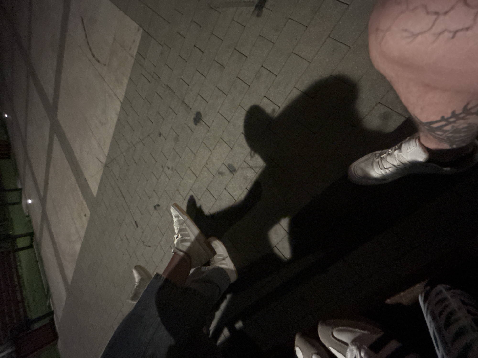 The image shows a low-angle view of several pairs of feet wearing white sneakers, with shadows cast on a paved area. The paving consists of rectangular grey stones arranged in a grid. In the background, a dark wooden bench and greenery are visible, suggesting an outdoor setting, possibly a park or a public seating area at dusk or night. The lighting creates distinct shadows of the people present, some of whom appear to be wearing jeans or patterned trousers. One individual's leg in the foreground exhibits visible veins and tattoo markings. There are no other discernible objects or activities in the immediate vicinity. The location of Siedlce, Poland, is not evident from the visual cues in the image.