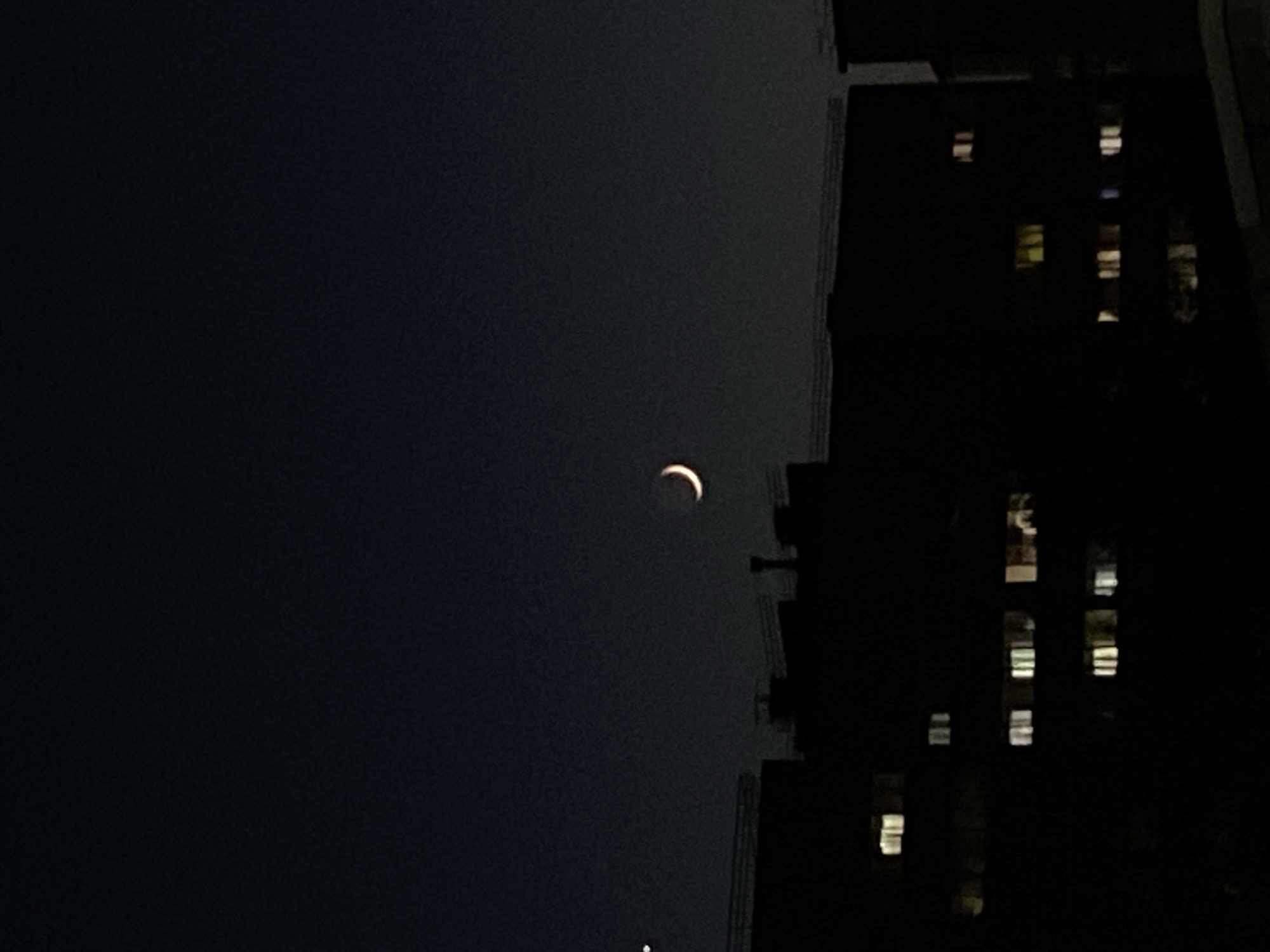 The image shows a crescent moon in the night sky. In the lower right, a dark building is visible with several illuminated windows. The exact location in Votkinsk, Russia is impossible to ascertain based on this image alone.