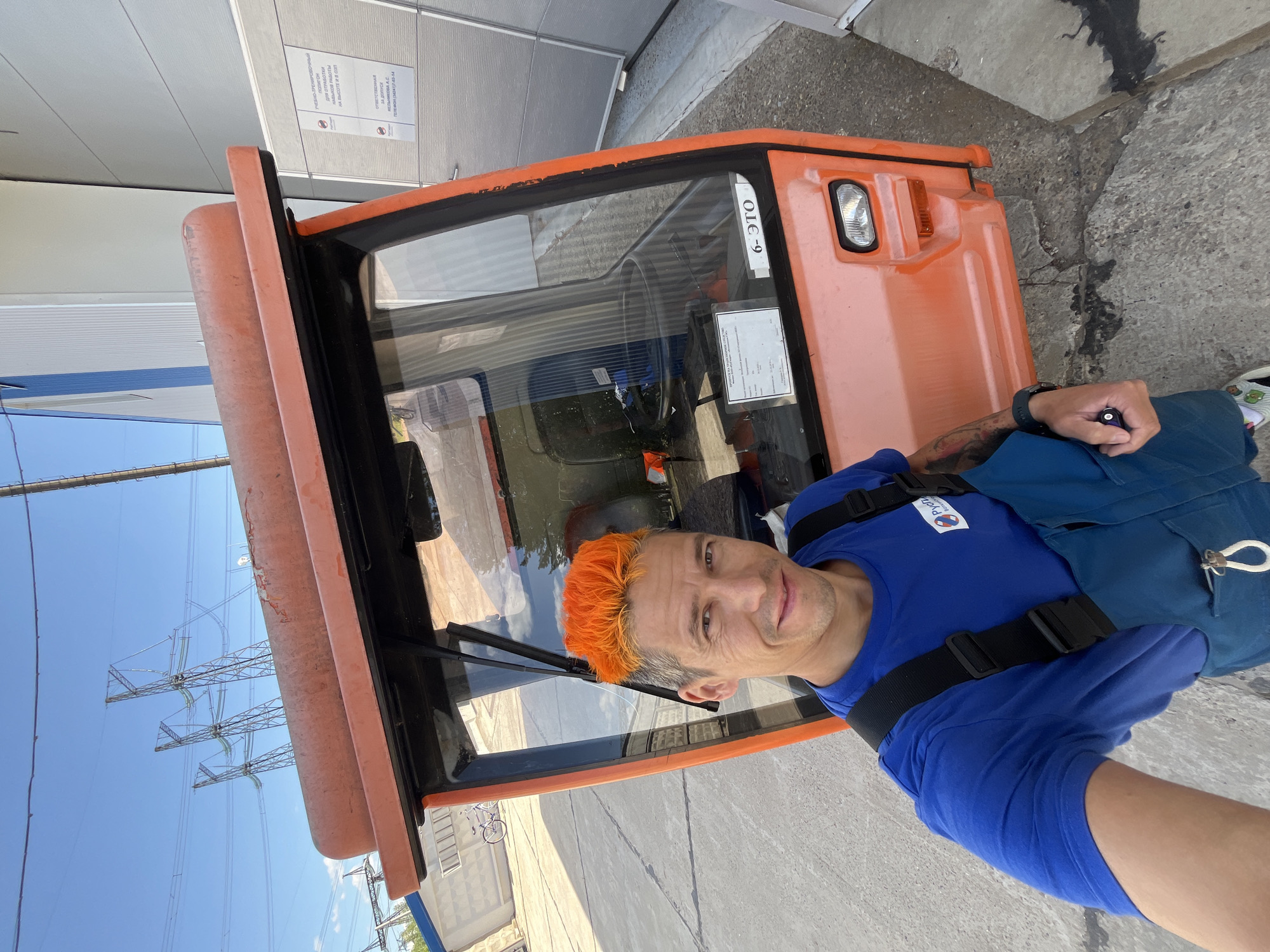 In an outdoor industrial setting, under a clear blue sky, a man with bright orange, spiky hair takes a selfie in front of an orange, boxy vehicle, possibly a utility vehicle or small truck. He is wearing a blue t-shirt and blue overalls. The vehicle has a glass cabin with a steering wheel visible inside. In the background, power lines and transmission towers are prominent, along with a modern building with large grey doors. A bicycle is visible in the distance. The ground is paved with concrete, showing some cracks and dark stains. The image appears to be taken in or around the city of Chaikovsky, Russia, suggested by a sign on the building in Russian.