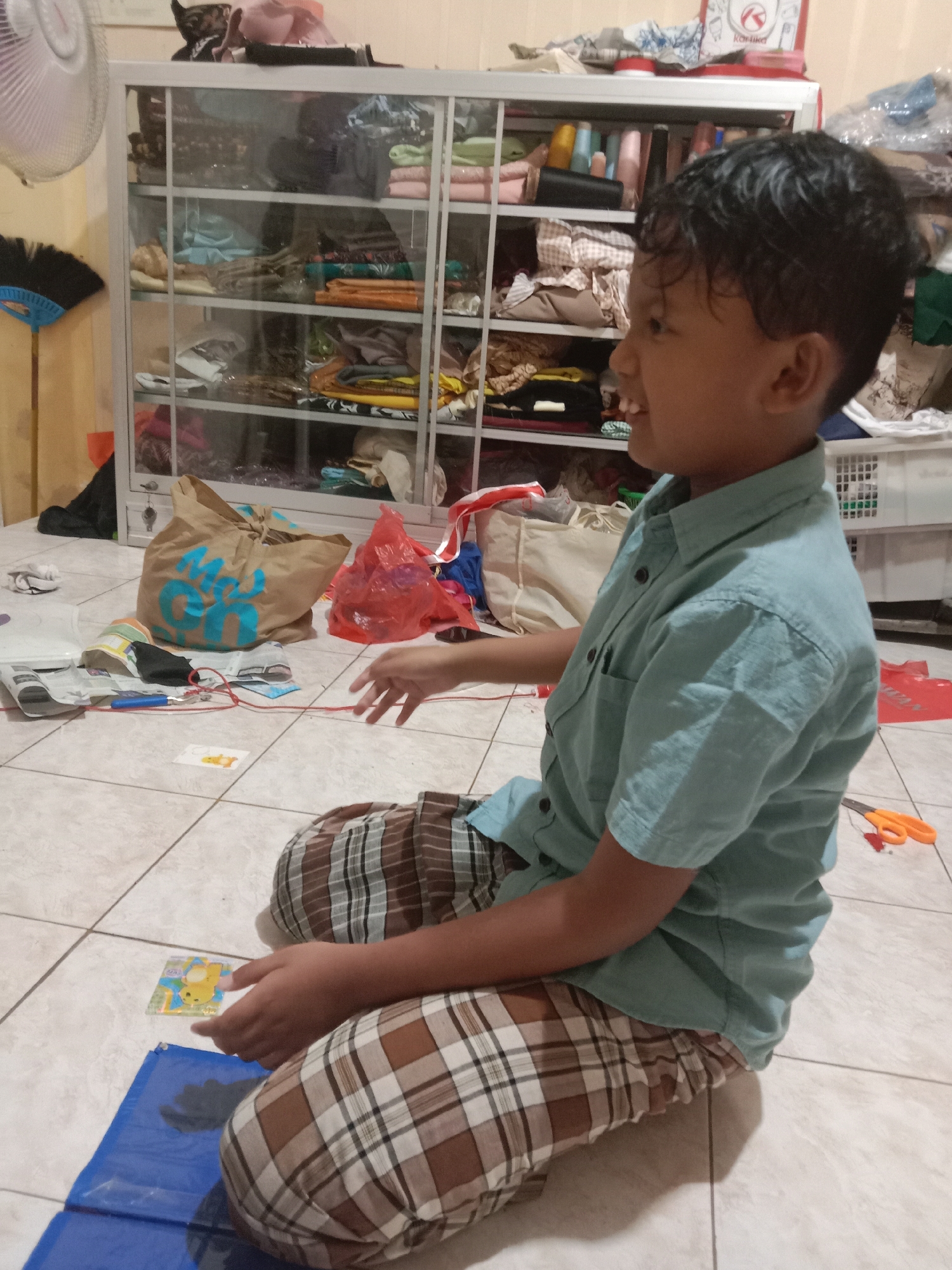 The image depicts a young boy sitting on a tiled floor, his side facing the camera. He is wearing a light blue collared shirt and plaid pants. The boy is looking to his right with his mouth open, appearing to be in mid-sentence or mid-action. In front of him, to his left, is a blue mat with a shadow on it and a small, yellow, rectangular object with an image on it. A pair of orange scissors lies on the floor to his right.
Behind the boy is a tall, glass-fronted cabinet filled with various folded items, possibly clothing or fabric. To the left of the cabinet, a part of a white fan is visible, along with a broom. Scattered on the floor in front of the cabinet are several bags and other items, including a brown paper bag with blue lettering and a red plastic bag. The overall environment suggests an indoor domestic setting, possibly a storage area or a room with stored goods. The lighting appears to be artificial, creating some shadows on the floor. There is no discernible indication of the specific location within Madiun, Indonesia.