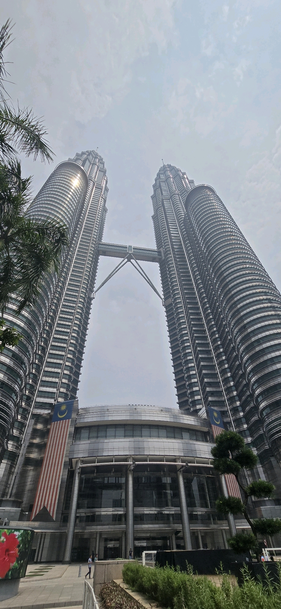 The image depicts the iconic Petronas Twin Towers in Kuala Lumpur, Malaysia, seen from a low-angle perspective looking upwards. The sky above is overcast with a hazy, pale blue color and scattered clouds.
The two skyscrapers, characterized by their distinctive metallic facade and fluted design, dominate the frame. A skybridge connects the two towers at a mid-level, with its supporting structures clearly visible. Malaysian flags are prominently displayed on the sides of the towers, near the base of each structure.
At the base of the towers, a modern, multi-story building with extensive glass windows and a metallic exterior is visible. The entrance area features a wide walkway with some landscaping, including trees and shrubs. A large digital display screen with an image of a red flower is present on the left side of the walkway.
Several individuals are present in the lower section of the image, walking on the paved area and near the entrance of the building. They are depicted as small figures, appearing to be tourists or visitors. The overall environment suggests an urban setting with contemporary architecture. The lighting is diffused due to the cloudy conditions.