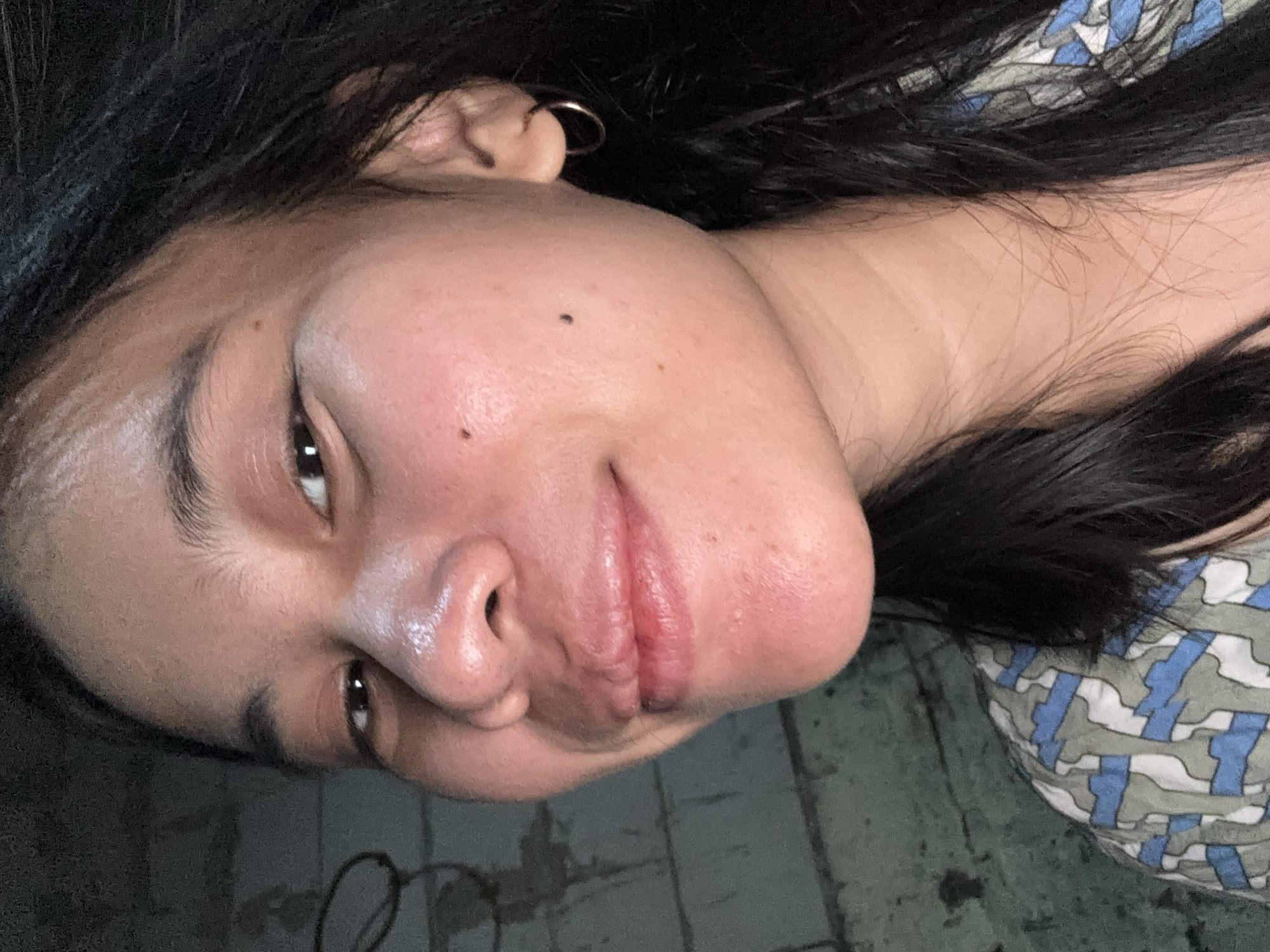 A close-up selfie captures a young woman with dark hair smiling faintly, her face angled slightly to the left. She wears a hoop earring in her right ear and a patterned shirt. Her skin appears to have a dewy sheen, and there are several moles visible on her face. The background consists of a textured, greenish-gray wall. The overall lighting is dim, with some highlight on her forehead and cheekbones. The image appears to have been taken indoors, potentially in a residential setting in East Jakarta, Indonesia.