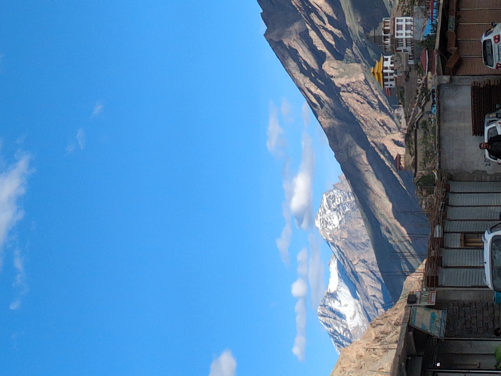 A vertical, eye-level shot captures the sprawling landscape and buildings in the city of Padum, India. The sky is a clear, bright blue with scattered white clouds. In the background, majestic, snow-capped mountains rise, their slopes casting long shadows under the sunlight. Nestled on the mountainside is a white monastery with a distinctive yellow roof and a stupa. In the foreground, the lower portion of the image shows a street scene with buildings made of corrugated metal and brick, some with faded signage. A white car is parked in front of one of the buildings, and a person can be seen standing near it. Another white car is visible further down the street. The overall impression is one of a remote and serene mountain town.