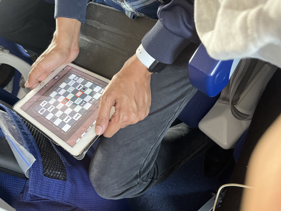 An overhead shot captures a person playing a digital chess game on a tablet, likely while seated on an airplane. The person's arms, hands, and legs are visible. The left hand holds the left side of the tablet, while the right hand rests near the right edge of the screen, with fingers extended. A white-banded smartwatch with a black face is worn on the left wrist. The person is wearing a dark blue long-sleeved top with ribbed cuffs and dark grey pants.
The tablet, possibly an iPad due to its design, has a white bezel and appears to be housed in a clear or light-colored protective case. The screen displays a digital chess board with standard white and black pieces. A black pawn on square d5 is highlighted with an orange-red outline, suggesting it is either selected or indicating a possible move. The chess board's alphanumeric coordinates (A-H, 1-8) are visible along its edges. Small numbers "1-8" and "8-1" are visible at the top of the screen, likely related to game statistics or captured pieces.
The setting appears to be an airplane cabin, indicated by the blue fabric of the seat, a dark grey surface resembling a tray table or armrest, and a prominent blue object that could be part of an armrest or seat fixture on the right. A seatbelt is also visible on the right. To the left, a clear plastic bag with a white zipper is tucked into a mesh pocket, likely on the side of the seat. The lighting is ambient, typical of an indoor environment. Although not visually confirmed in the image, the provided context indicates this scene takes place in Guwahati, India.
