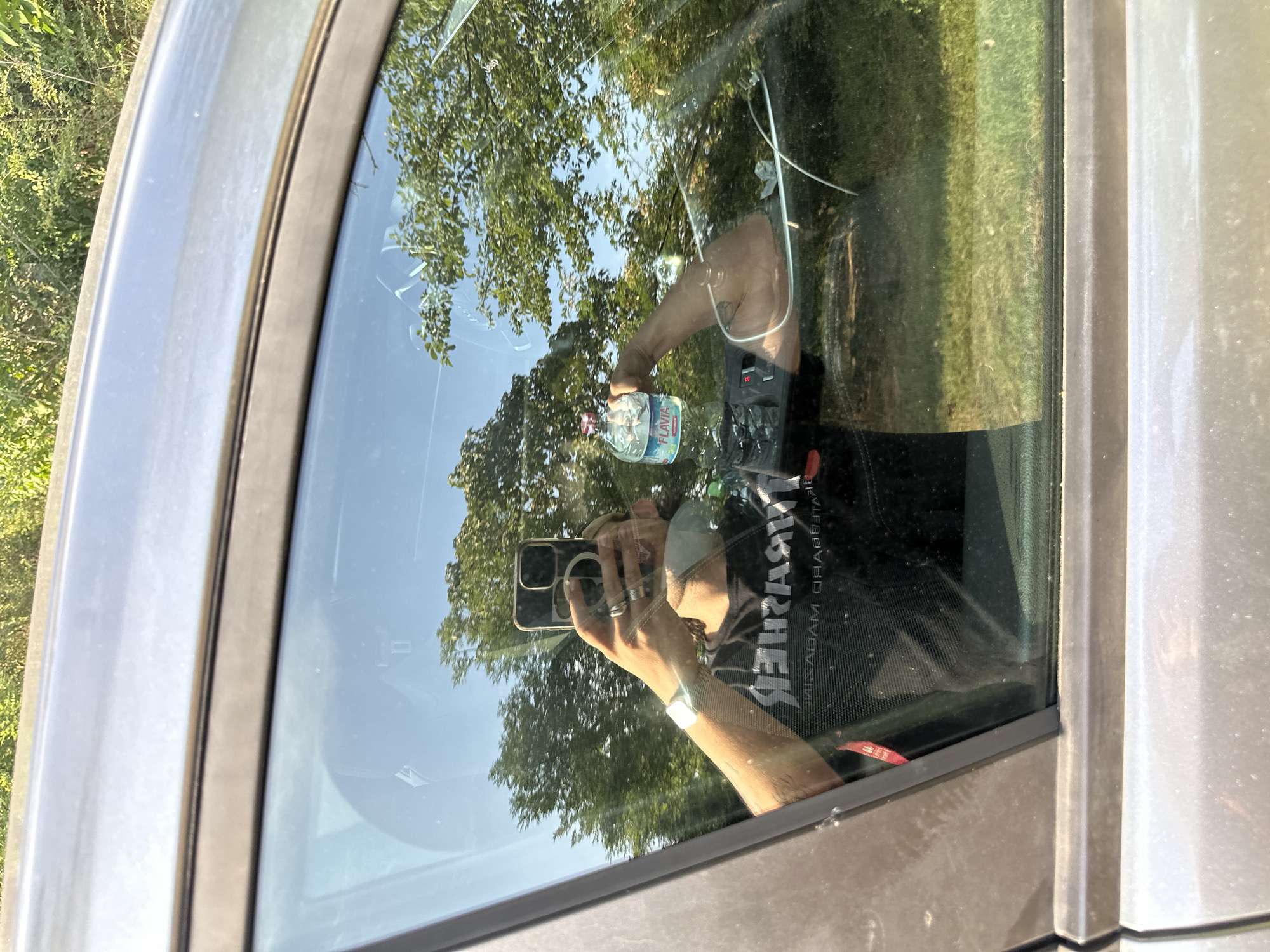 In Rudiano, Italy, a person is reflected in a car window, holding a smartphone and a bottle of "Flavia" brand water. The individual is wearing a black t-shirt with white lettering that reads "THRASHER" in reverse. They have a watch on their left wrist and several rings on their fingers. The car's interior is visible in the reflection, including a portion of the dashboard. Outside the car, trees and a bright blue sky are visible. The car appears to be parked in an outdoor setting, possibly a parking lot or roadside, given the presence of greenery and natural light. The image is taken from a low angle, focusing on the reflection in the window.