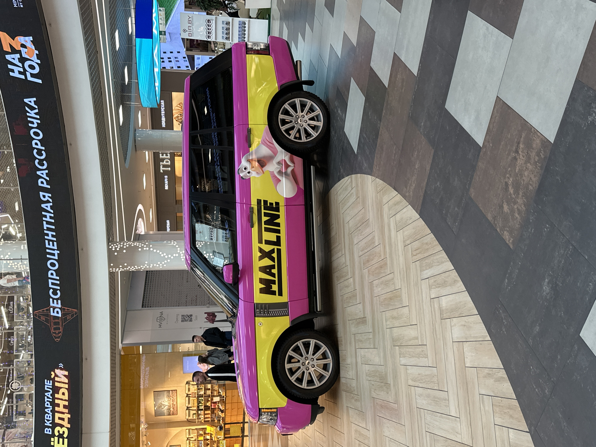 In an indoor shopping mall, a customized pink and yellow Range Rover is parked on a herringbone-patterned floor. The vehicle is decorated with the text "MAXLINE" and an image of a cartoon duck wearing a pink shirt and making a heart shape with its hands. The mall ceiling features a large digital display advertising "3 YEARS", "INSTALMENT-FREE", and "FOR THE YEAR". In the background, several shops are visible, including "MARCO OZZI", "Tamaris", "Cath", and "s.Oliver", along with people walking. The floor transitions to a dark tiled pattern with rectangular tiles of varying shades.