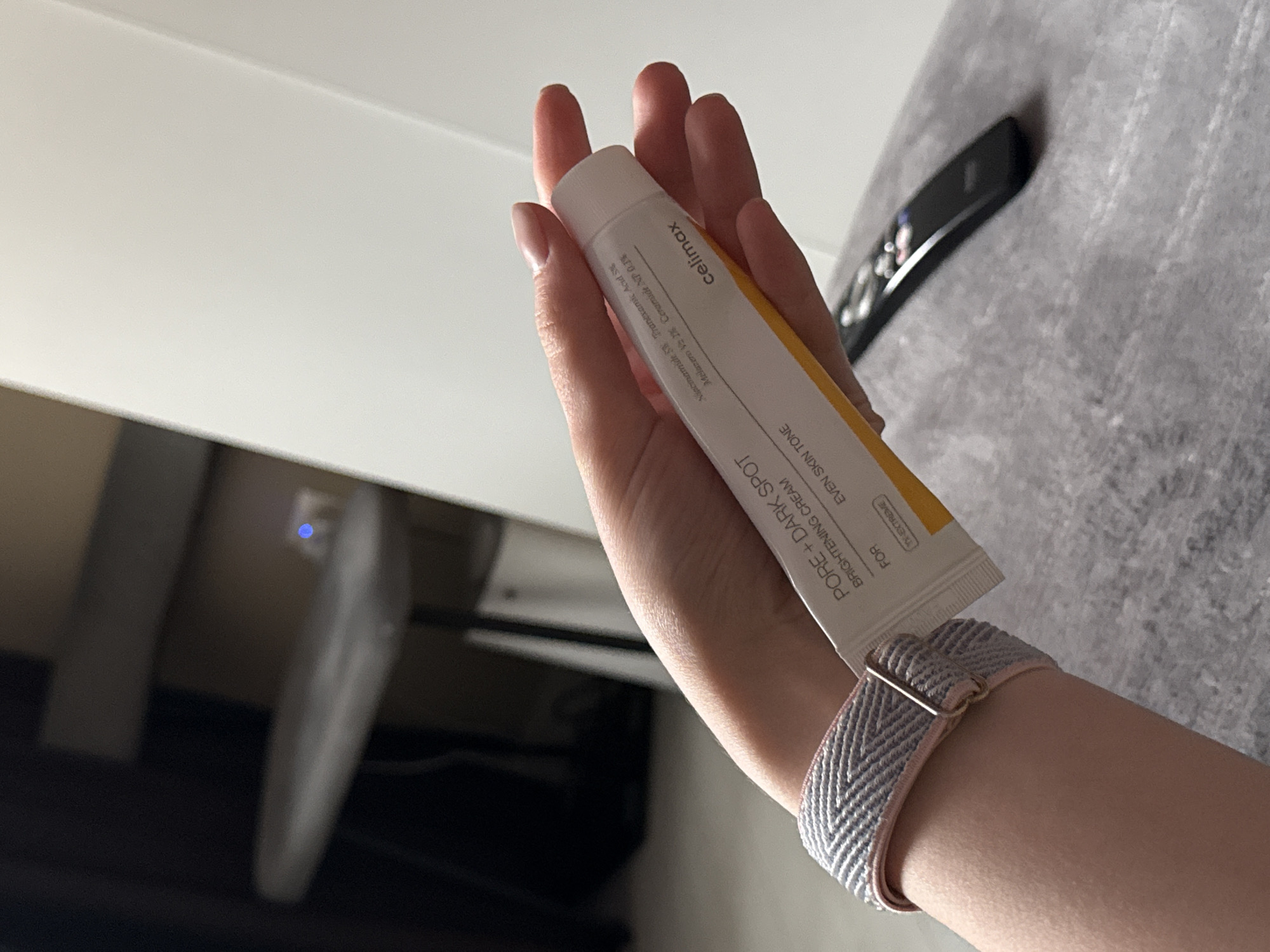 A hand holds a tube of "celimax PORE + DARK SPOT BRIGHTENING CREAM FOR EVEN SKIN TONE" in front of a background featuring a gray-toned surface and a television on a stand. The hand is fair-skinned with a gray and pink woven watch band on the wrist. The cream tube is white with yellow accents and contains ingredients such as "Niacinamide 5% Tranexamic Acid 5%" and "Melazero V2 1% Ceramide NP 0.1%." The background consists of a gray, textured surface, possibly a countertop or table, and a blurred television screen and stand.