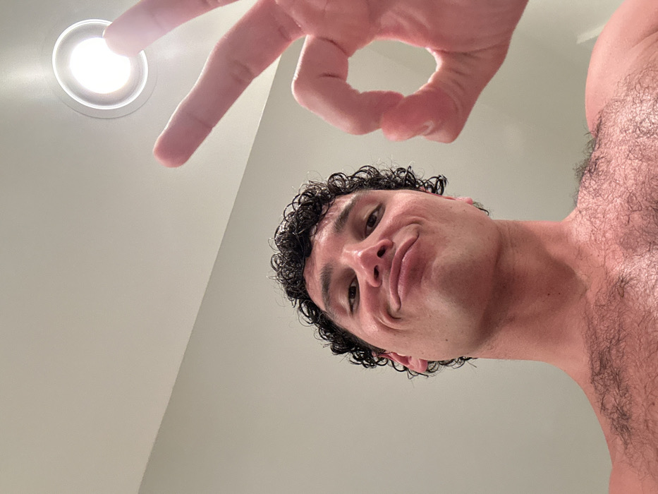 The image shows a man with curly brown hair, making an "ok" sign with his right hand towards the camera. He is wearing no shirt and has a light brown complexion with light hair on his chest. The man's facial expression suggests he is smiling slightly. The background is a pale green wall with a white ceiling. There is a bright, white, circular light fixture in the ceiling to the right. The image appears to be a selfie taken in a bathroom or a room with a bathroom-like appearance, at 25.773 latitude and -80.14 longitude, located in Miami Beach, Florida, United States.
