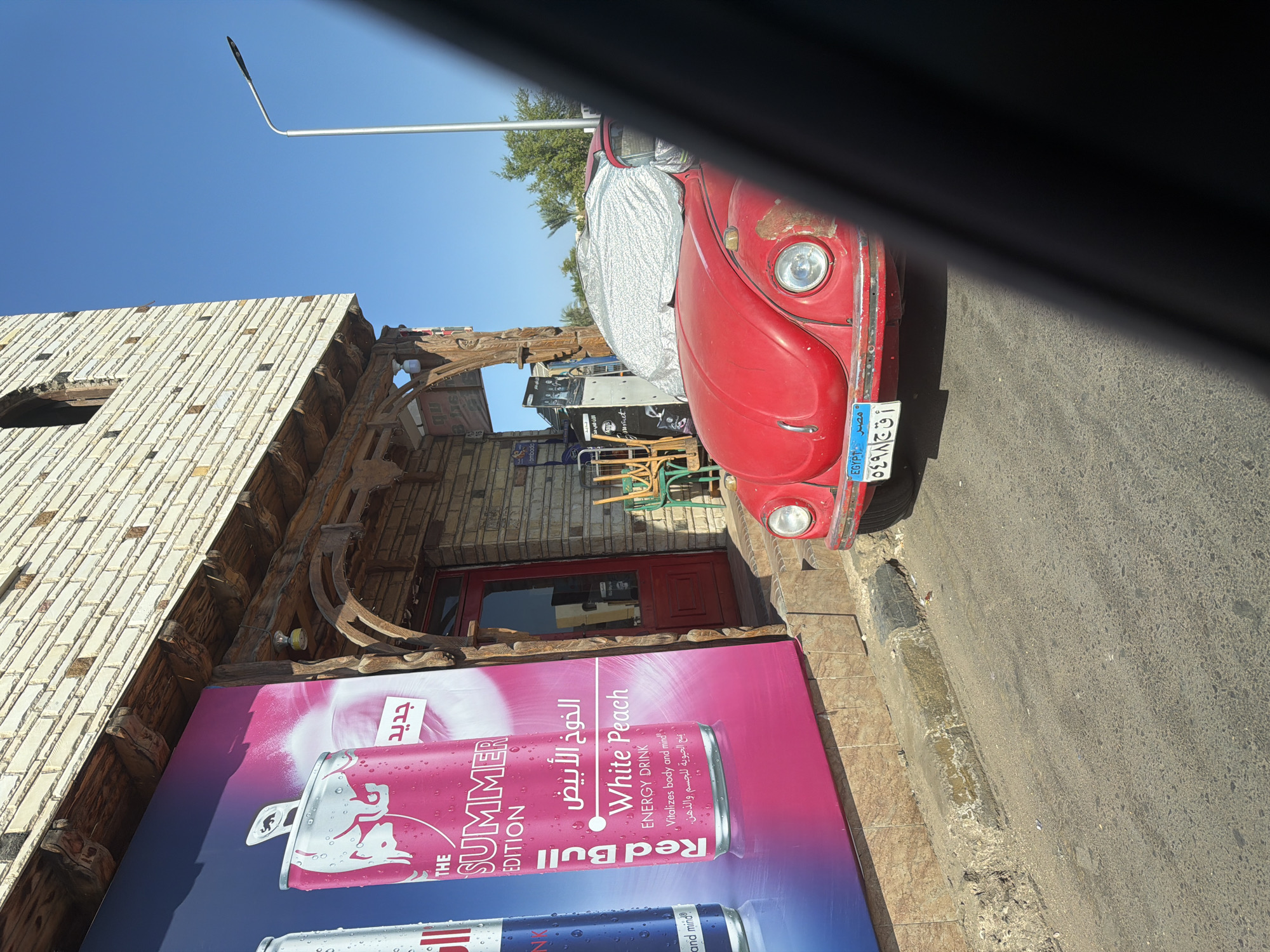 The image depicts a street scene in Dahab, Egypt, dominated by a vibrant Red Bull advertising billboard to the left. The billboard features a large can of Red Bull's "Summer Edition" in White Peach flavor, with Arabic text above the English description. Adjacent to the billboard is a building constructed with textured stone and wood, featuring a red door and arched wooden architectural elements. In front of the building, a classic red Volkswagen Beetle is parked, partially covered with a reflective car cover. The car has an Egyptian license plate that reads "مصر EGYPT" followed by alphanumeric characters. Green chairs are stacked near the entrance of the building, and a street light is visible in the background against a clear blue sky. The street in the foreground is asphalt, with a raised concrete curb. The overall impression is a snapshot of a roadside business in a sunny, possibly warm, environment.