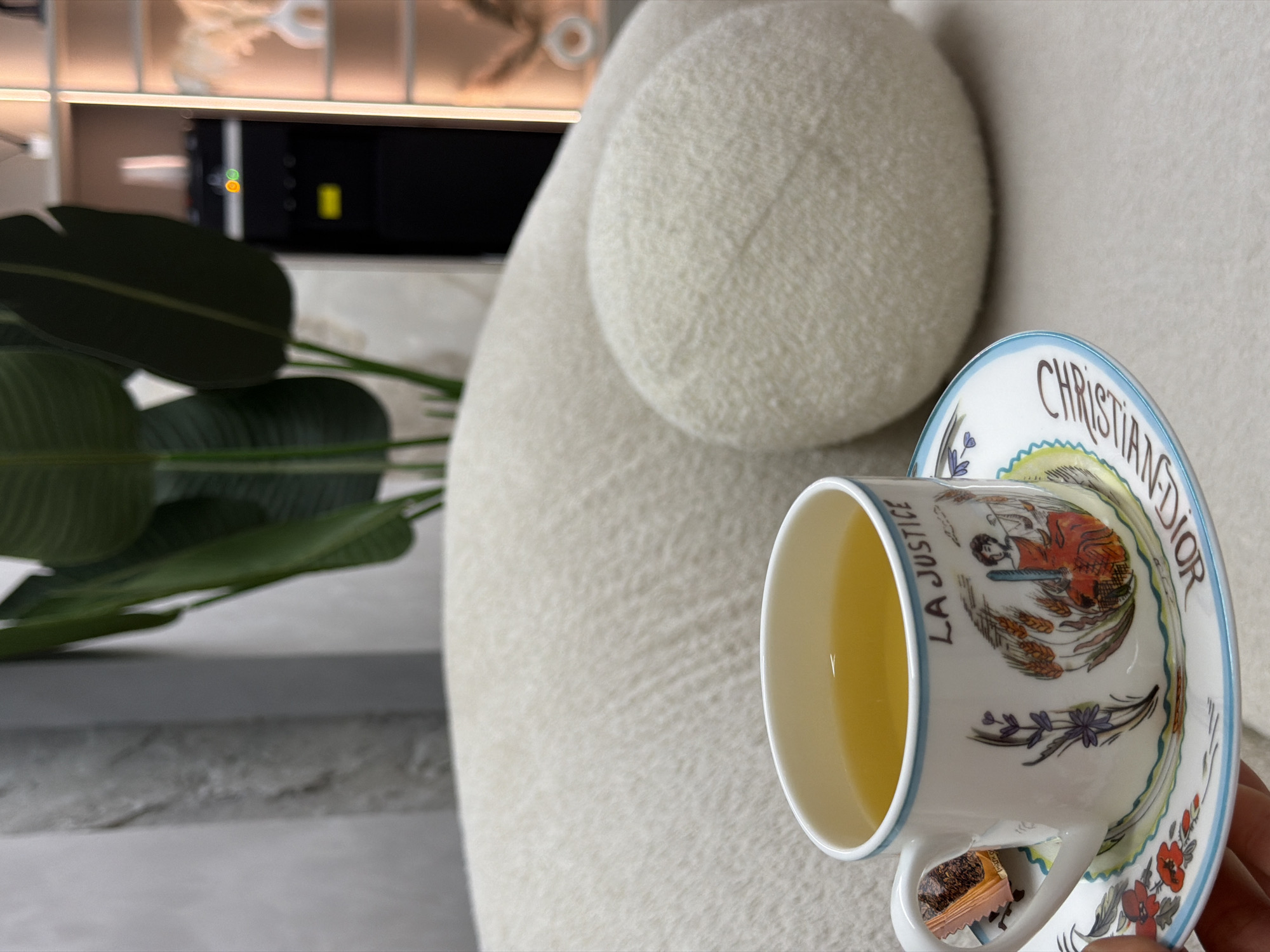 A teacup and saucer with "Christian Dior" and "La Justice" printed on them sit next to a large, white, fuzzy couch, with a house plant in the background. There is no evidence to suggest this photo was taken in Krasnoyarsk, Russia.