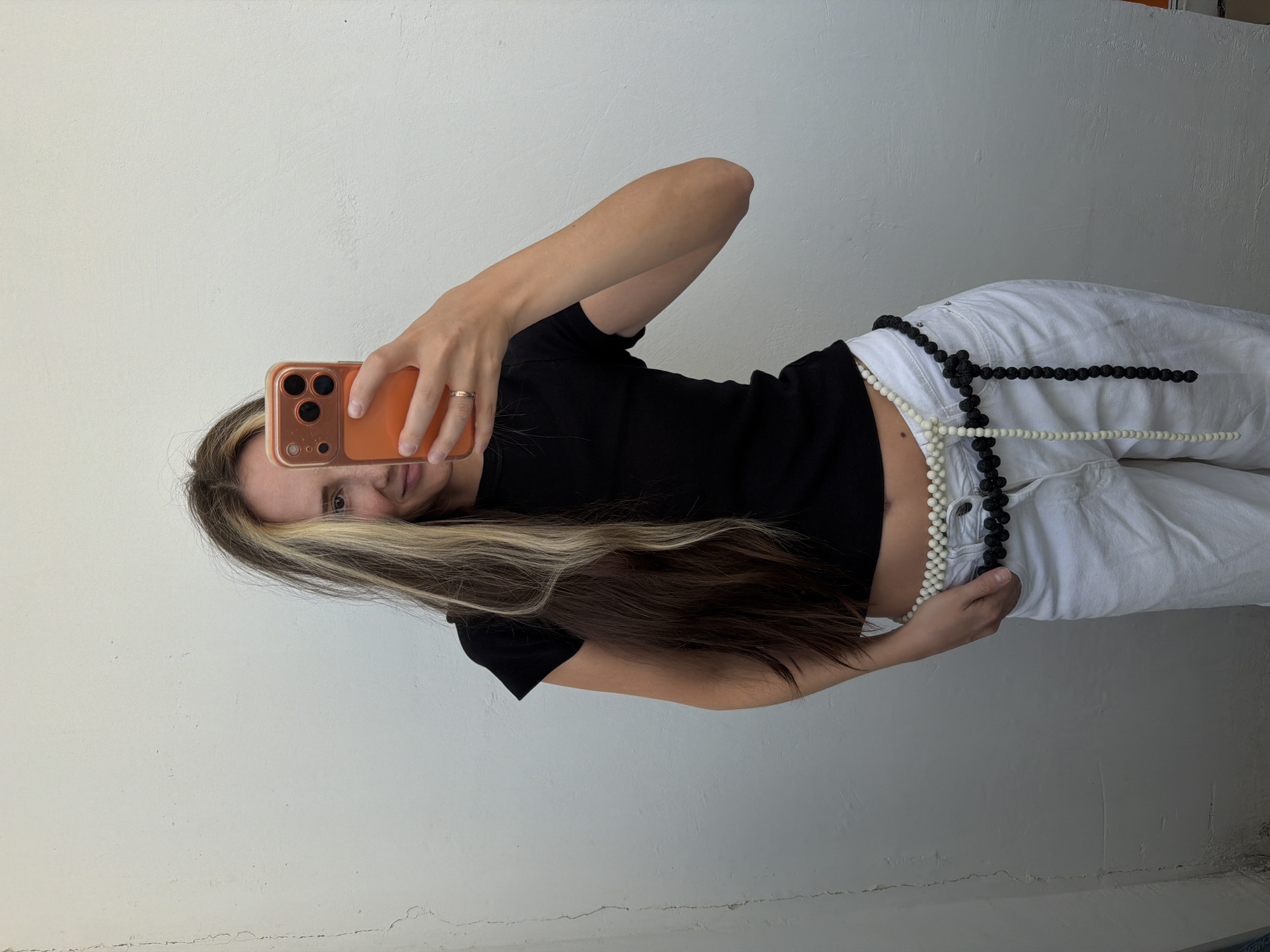 A woman with long, multi-colored hair, a black t-shirt, and white pants is taking a selfie against a textured, light-colored wall. She is holding an orange smartphone in her right hand, which is covering most of her face. She is wearing a beaded belt, with strands of white and black beads. The wall shows signs of age and weathering, with visible cracks and an uneven surface. The scene appears to be outdoors in an urban setting, possibly a courtyard or alleyway, in Khimki, Russia.