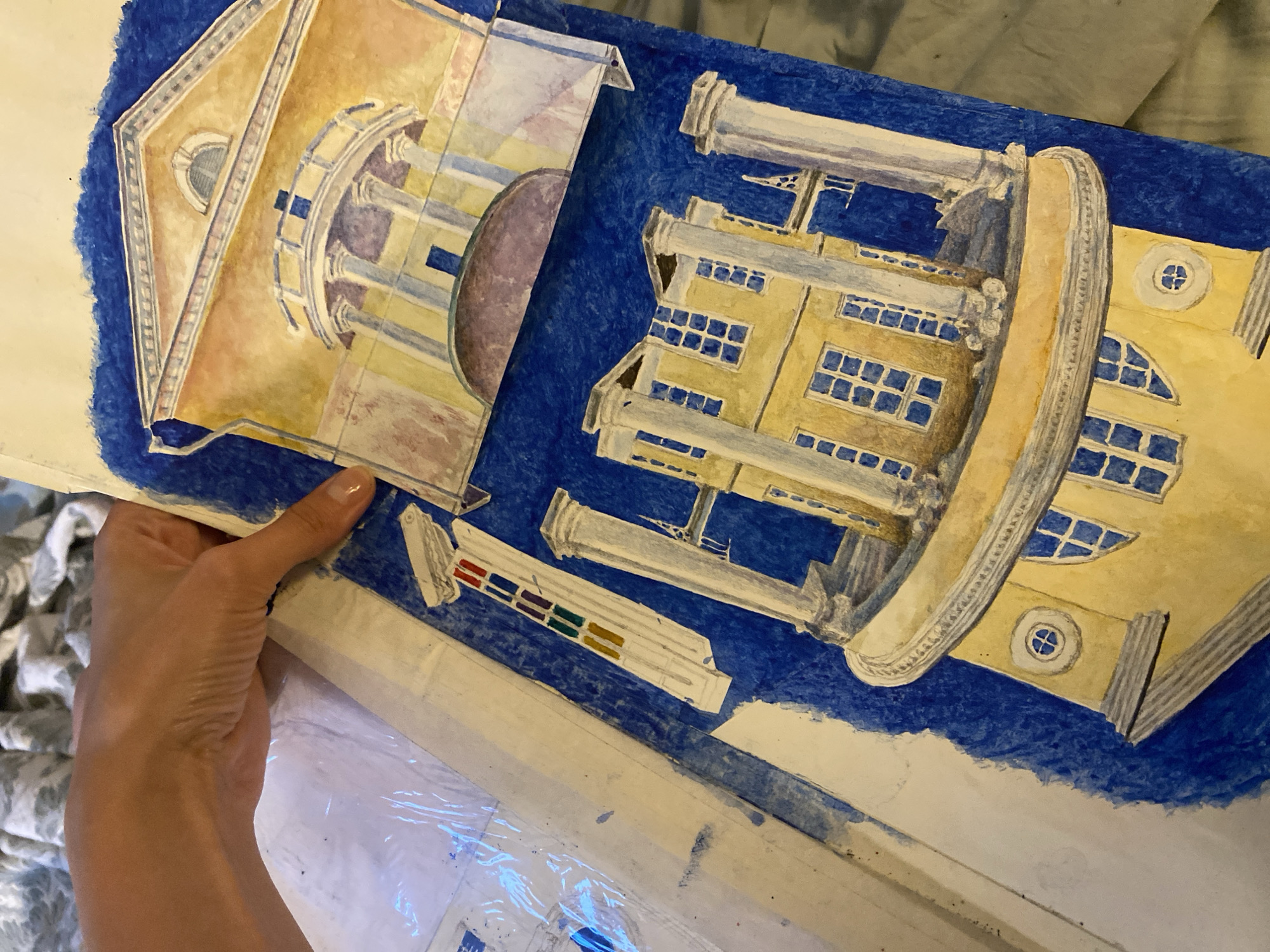 The image shows a close-up of a watercolor painting of architectural elements, specifically a facade with columns and windows. A person's hand is visible on the upper left, holding the painting. The painting is on blue paper, with sections of the artwork appearing to be layered or attached with tape. One section depicts a building with a curved balcony supported by columns, rendered in yellow and white with blue window details. Another section, positioned above the first, shows a similar architectural style with a triangular pediment and a semicircular window. The style suggests classical architecture, possibly representing a building in Saint Petersburg, Russia, given the prompt's context.