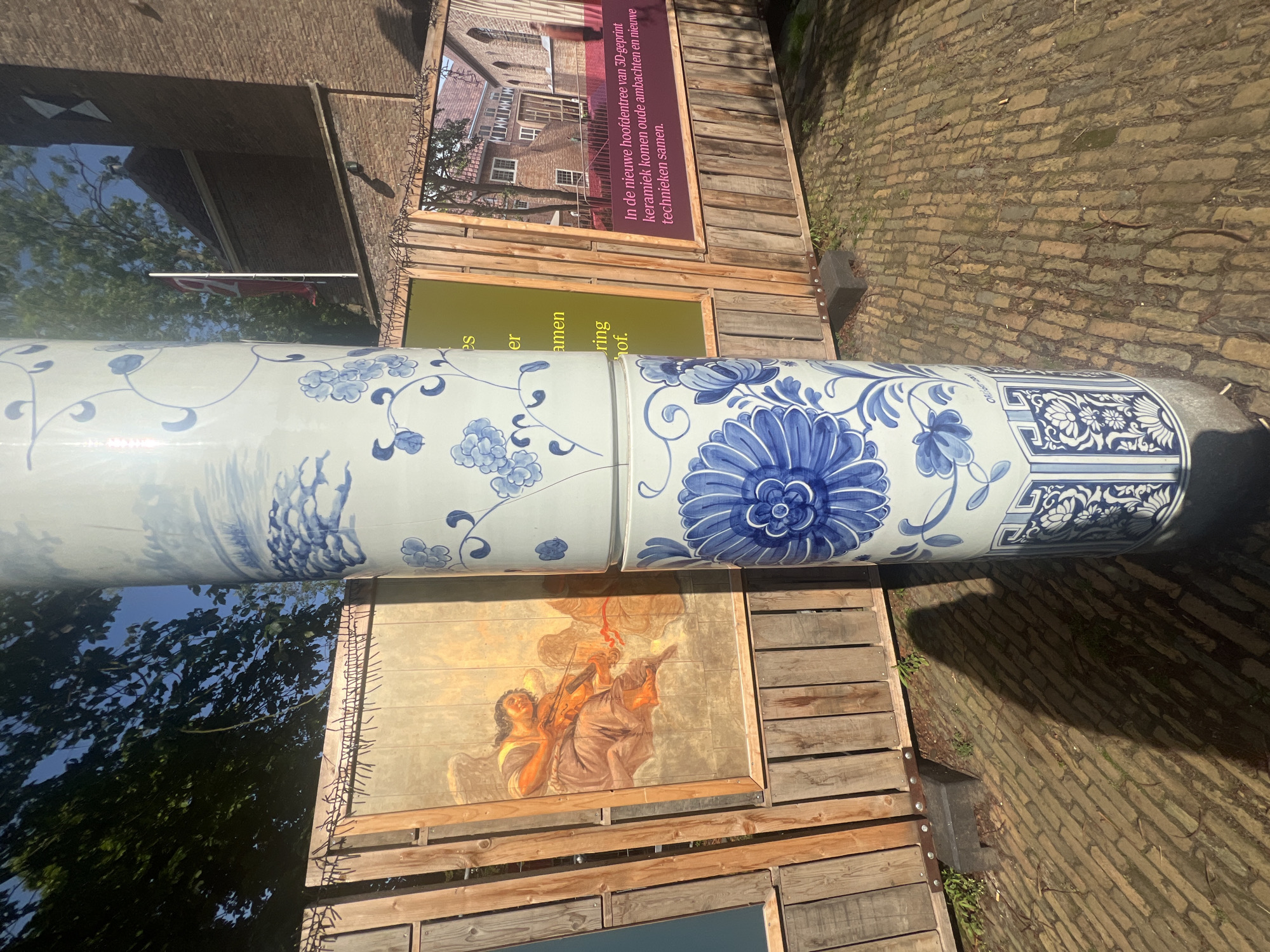 The image captures an outdoor scene in Delft, Netherlands, featuring two stacked cylindrical ceramic pillars decorated with blue and white Delftware patterns. The pillars stand on a brick-paved ground. Behind the pillars, a wooden fence structure is visible, displaying various artworks and informational boards.
On the left, a large framed artwork depicts an angelic figure playing a violin. To the right of the central pillars, another framed piece shows a photograph of a building, possibly a historical Dutch building, with text in Dutch printed on a purple overlay. The text reads: "In de nieuwe hoofdentree van 3D-geprint keramiek komen oude ambachten en nieuwe technieken samen." which translates to: "In the new entrance of 3D-printed ceramics, old crafts and new techniques come together." A red flag on a flagpole is also visible in the background, with a church spire and brick buildings. The scene is illuminated by sunlight, casting shadows on the ground.
