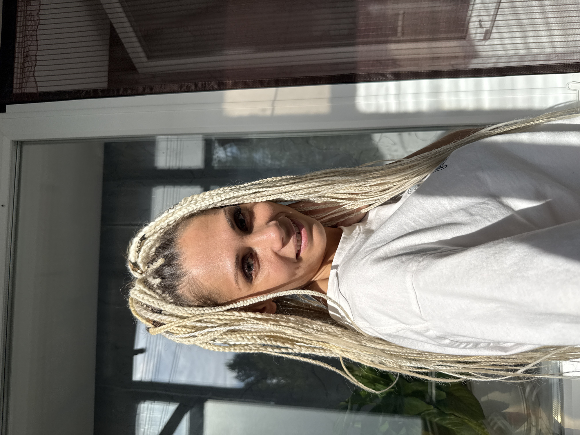 A medium shot captures a woman with long, blonde, braided hair smiling at the camera. She is wearing a white t-shirt and is positioned in front of a large window. Sunlight streams in from the right, casting shadows across the scene. Outside the window, a glimpse of greenery and sky is visible. To the right of the window, a sheer, dark-colored curtain with vertical stripes is present. The overall impression is that of a casual, outdoor portrait, possibly taken during the daytime in a residential setting. The location of Rusanovo, Russia is not directly depicted in the image but is provided as context.